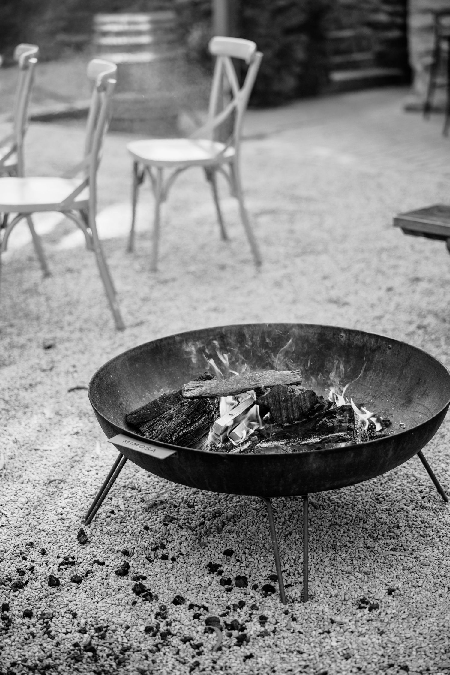 Black and white photo of a cast iron fire pit with burning logs on a patio with white chairs in the background.