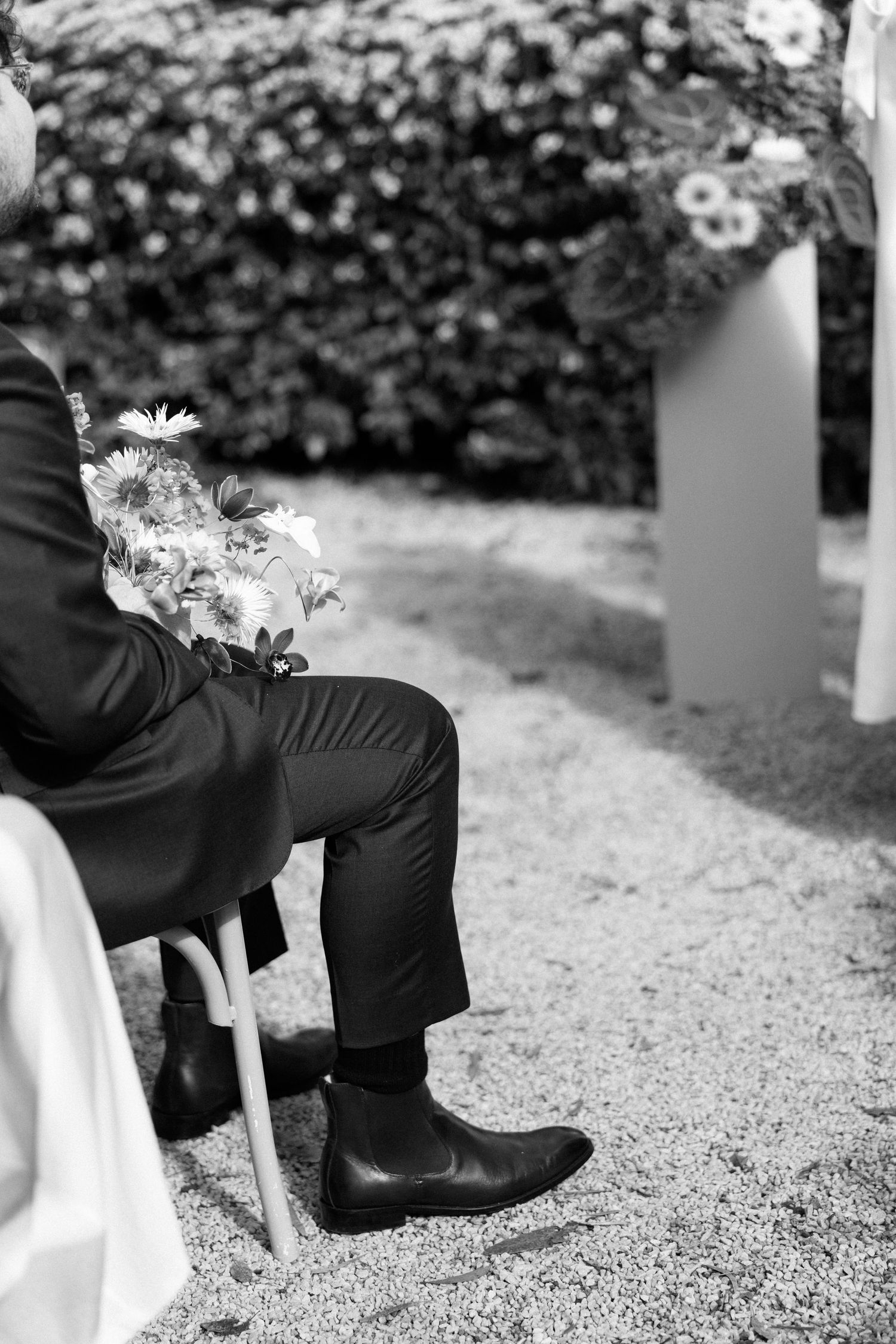 Black and white photo of a groom in suit and Chelsea boots seated outdoors at a wedding ceremony with flowers.