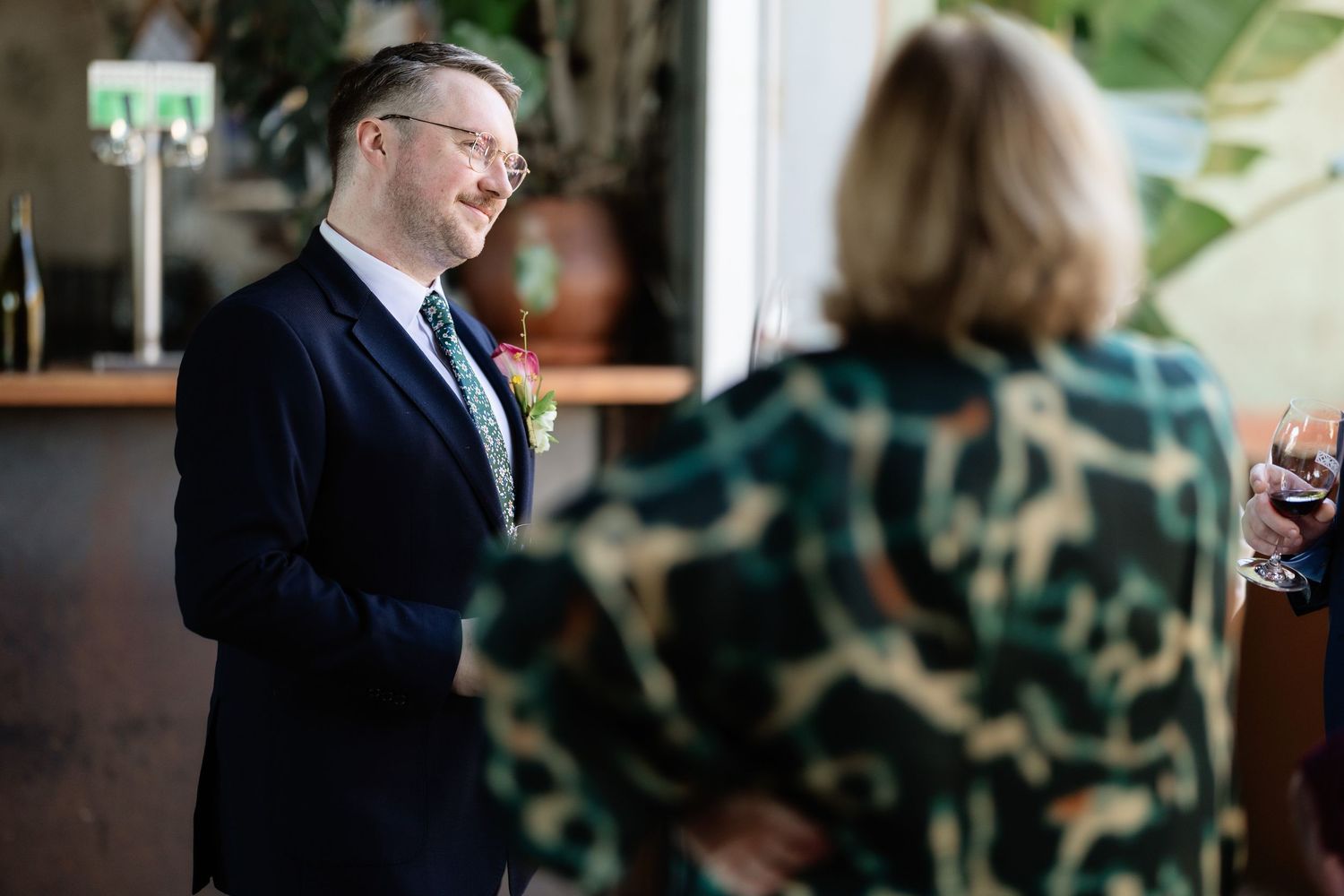 Groom in navy suit smiling at guest during wedding reception in elegant venue.