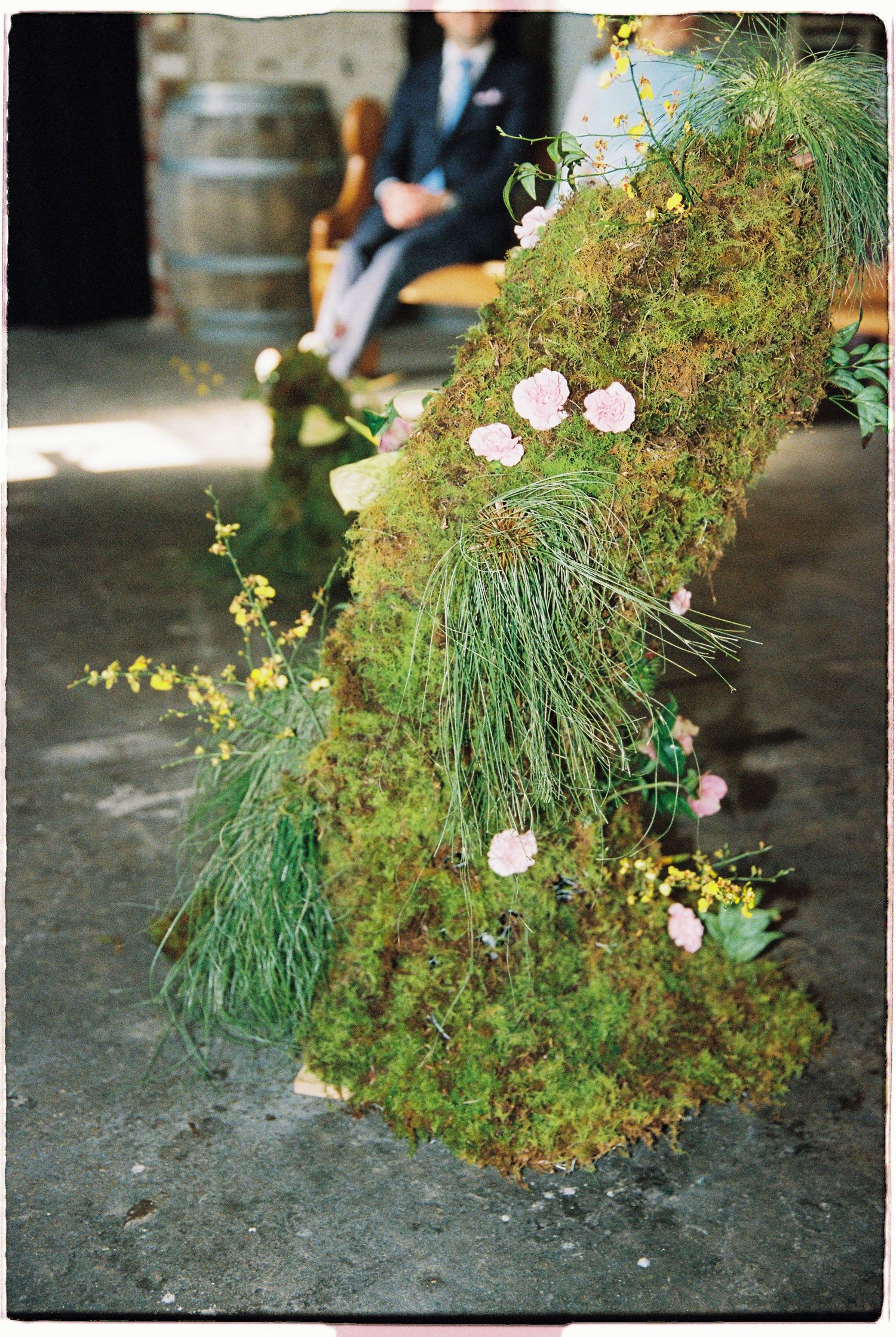 Moss-covered wedding arch adorned with pink roses and yellow wildflowers at a rustic winery venue.