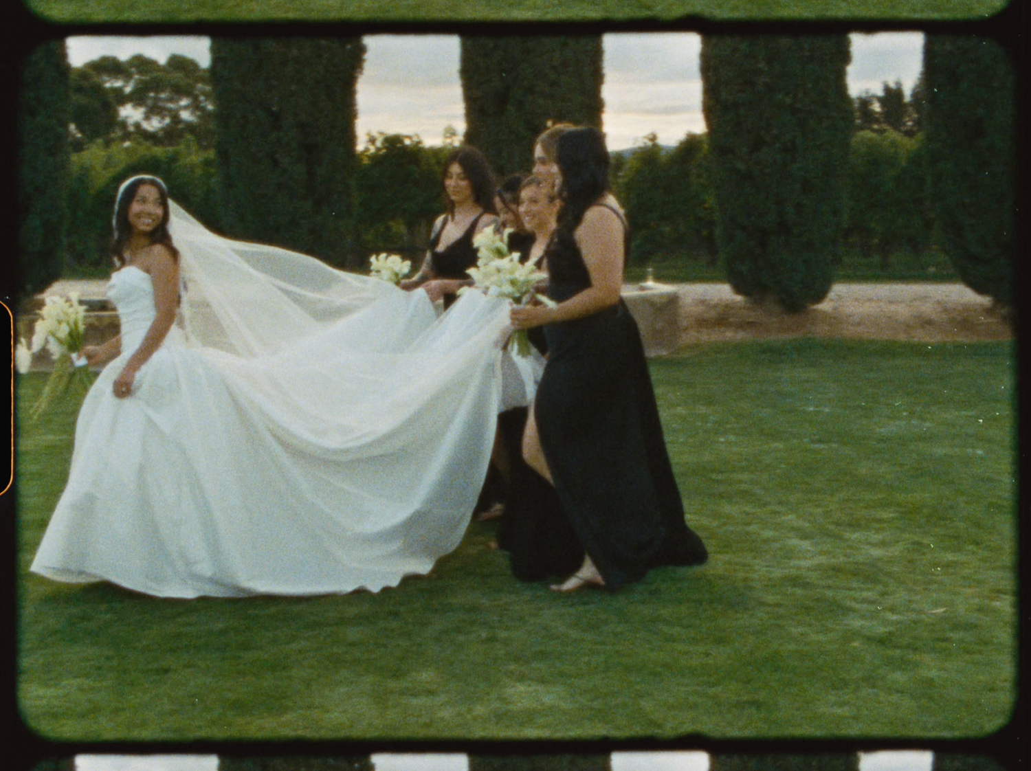 Bridesmaids in black dresses help bride with long white wedding gown train on green lawn.