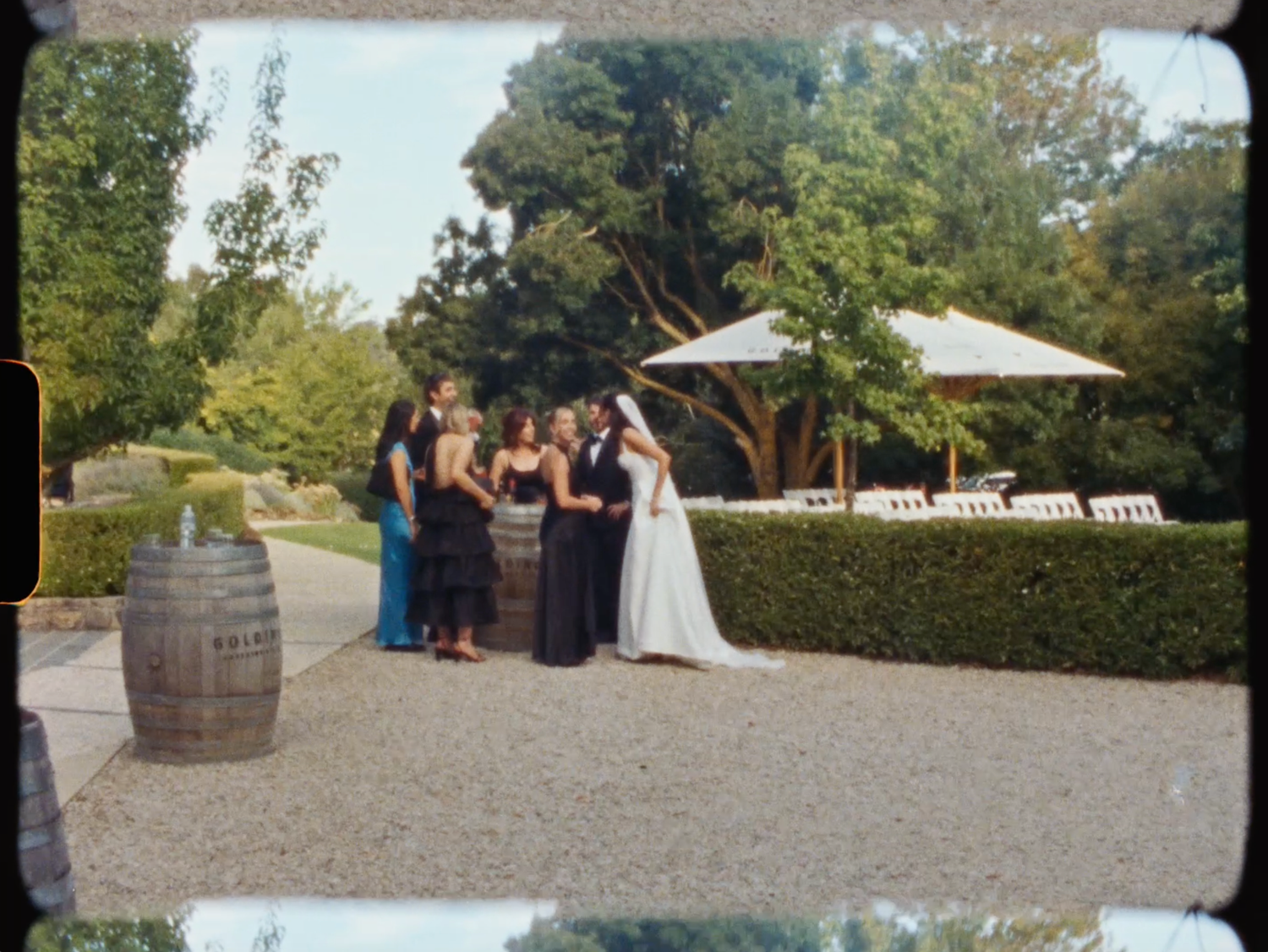 Wedding party gathers near a wooden barrel in a scenic garden venue with tall hedges and umbrella.