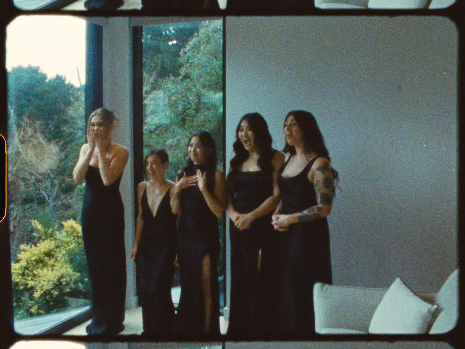 Five bridesmaids in black gowns stand together near floor-to-ceiling windows during a wedding getting-ready moment.