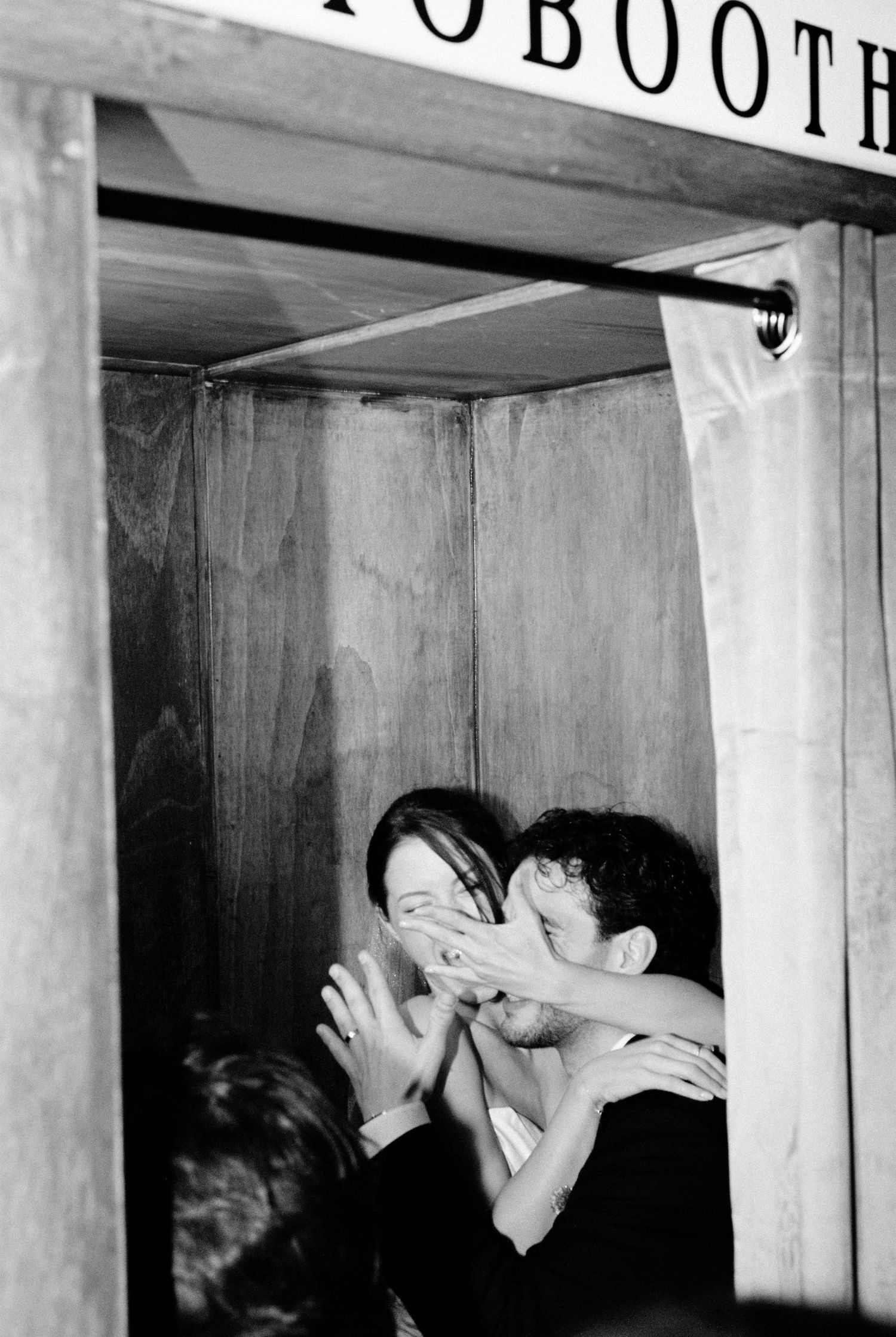 Couple playfully covering faces inside a vintage photo booth in black and white.