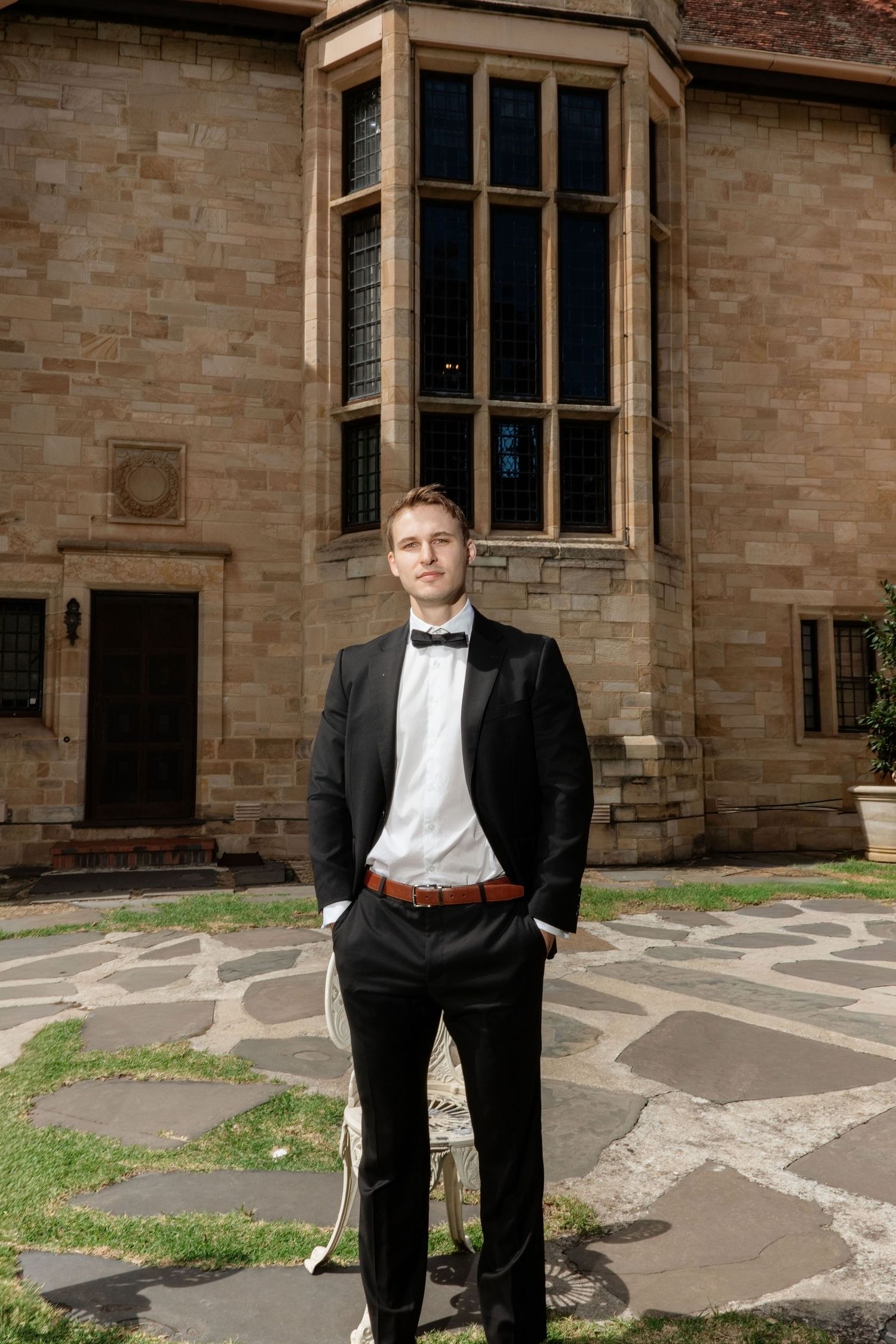 Dapper groom in black tuxedo with bow tie standing confidently outside historic sandstone building on wedding day.