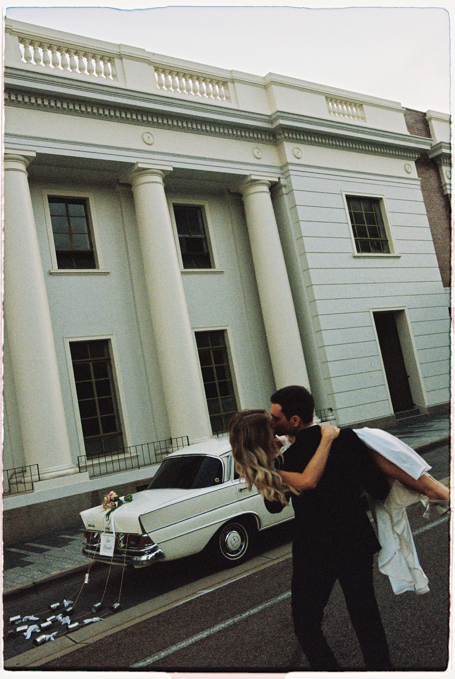 Groom lifting bride in a kiss outside white neoclassical building with vintage wedding car.