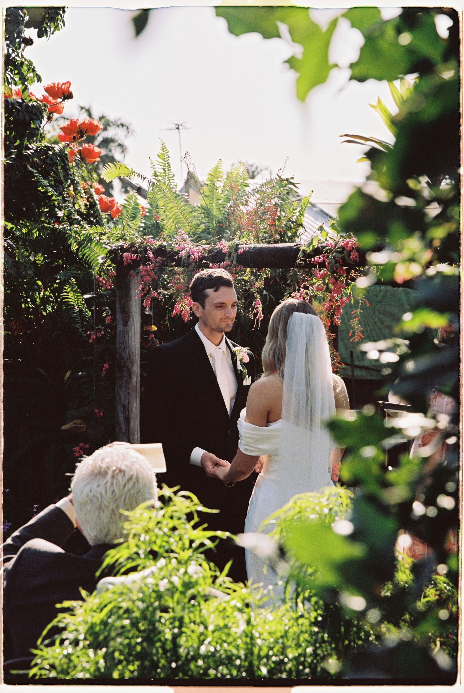 Groom in black suit exchanges vows with veiled bride under floral arch surrounded by lush garden greenery.