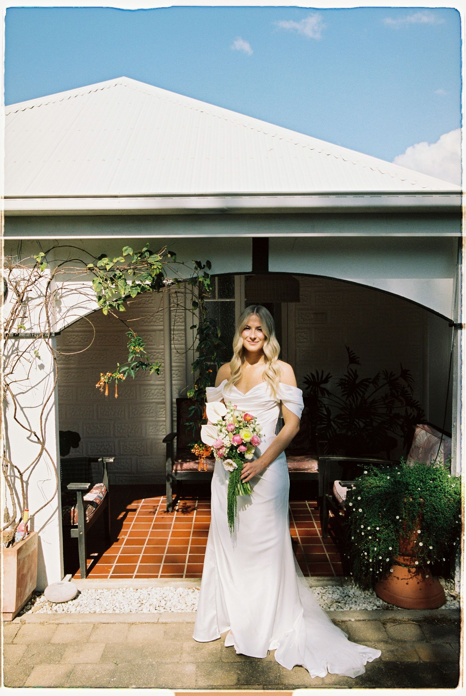 Bride in off-shoulder white gown holding colorful bouquet standing at entrance of home.
