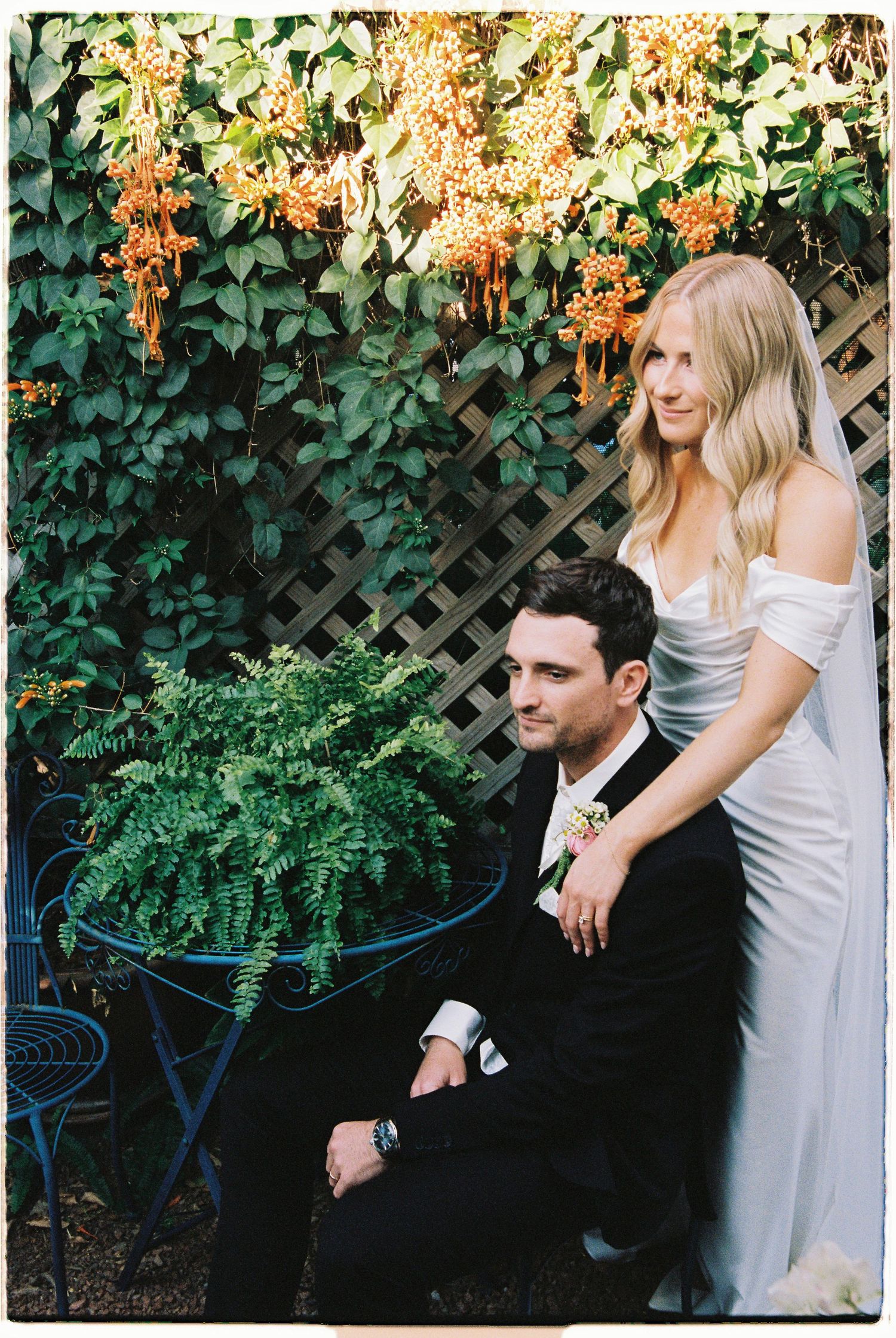 Bride in off-shoulder gown stands behind groom in tuxedo seated outdoors near lush green ivy wall with orange blooms.