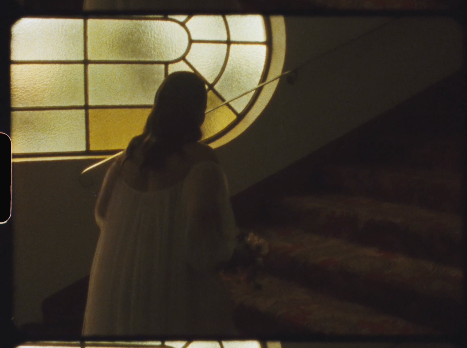 A woman in a white dress stands before a stained glass window on a staircase in soft golden light.