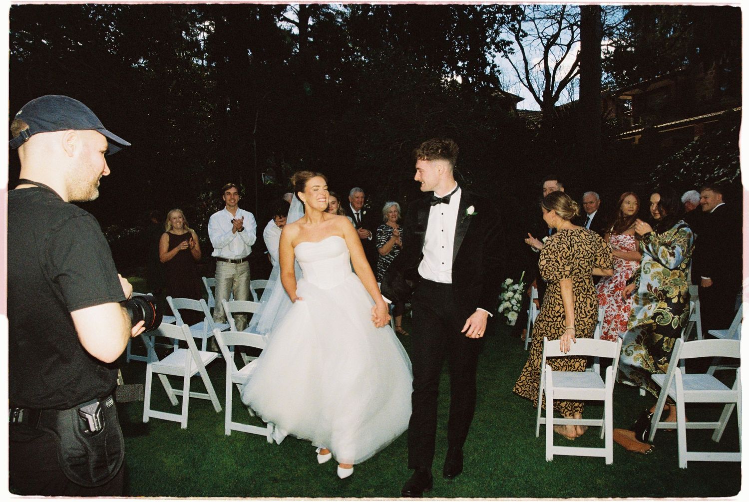 Bride and groom walk joyfully down aisle after ceremony as guests applaud in outdoor wedding.