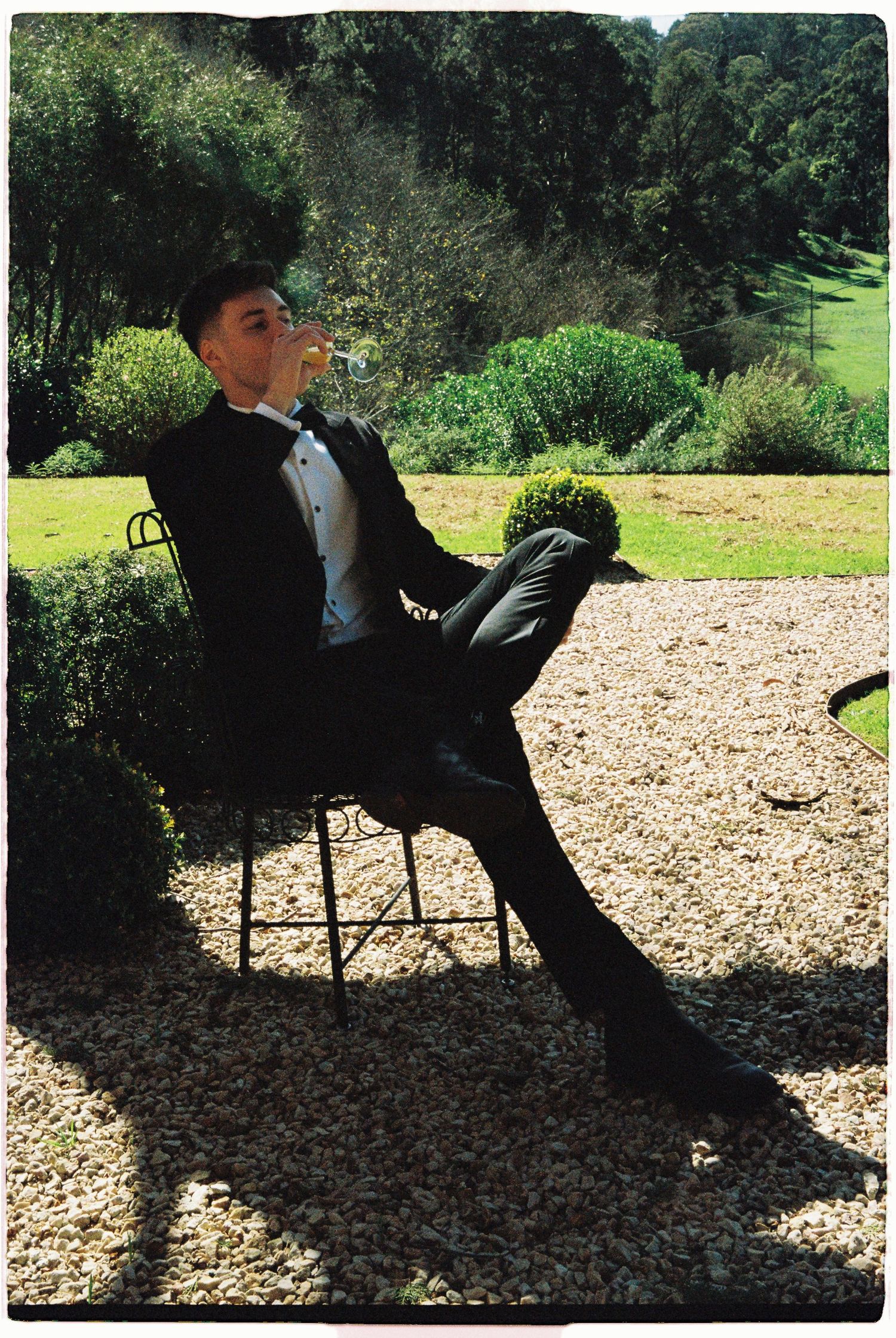 Groom in black tuxedo sitting in garden chair sipping champagne on sunny gravel terrace.