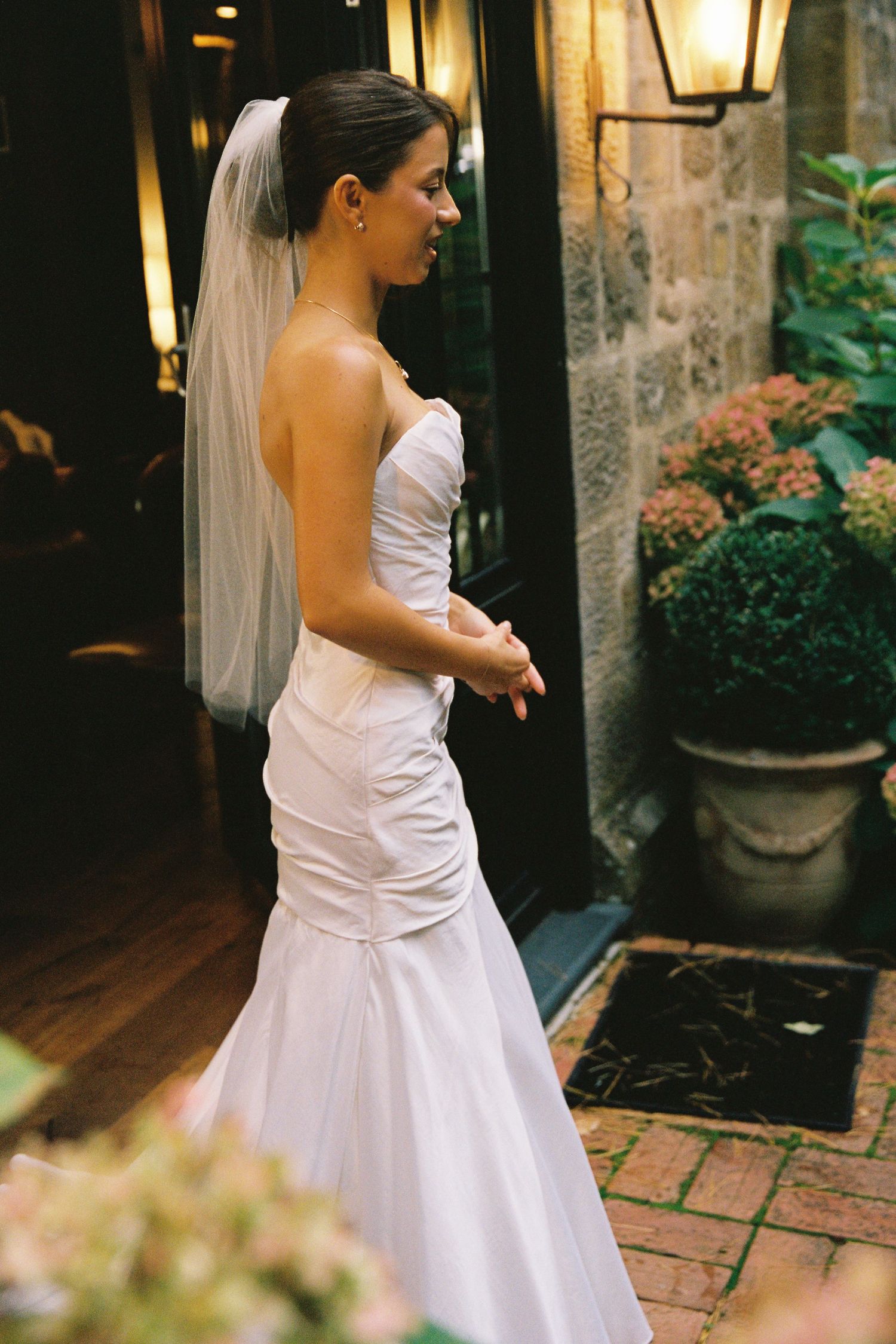 Elegant bride in a strapless mermaid wedding gown with veil standing outdoors near stone walls and flowers.