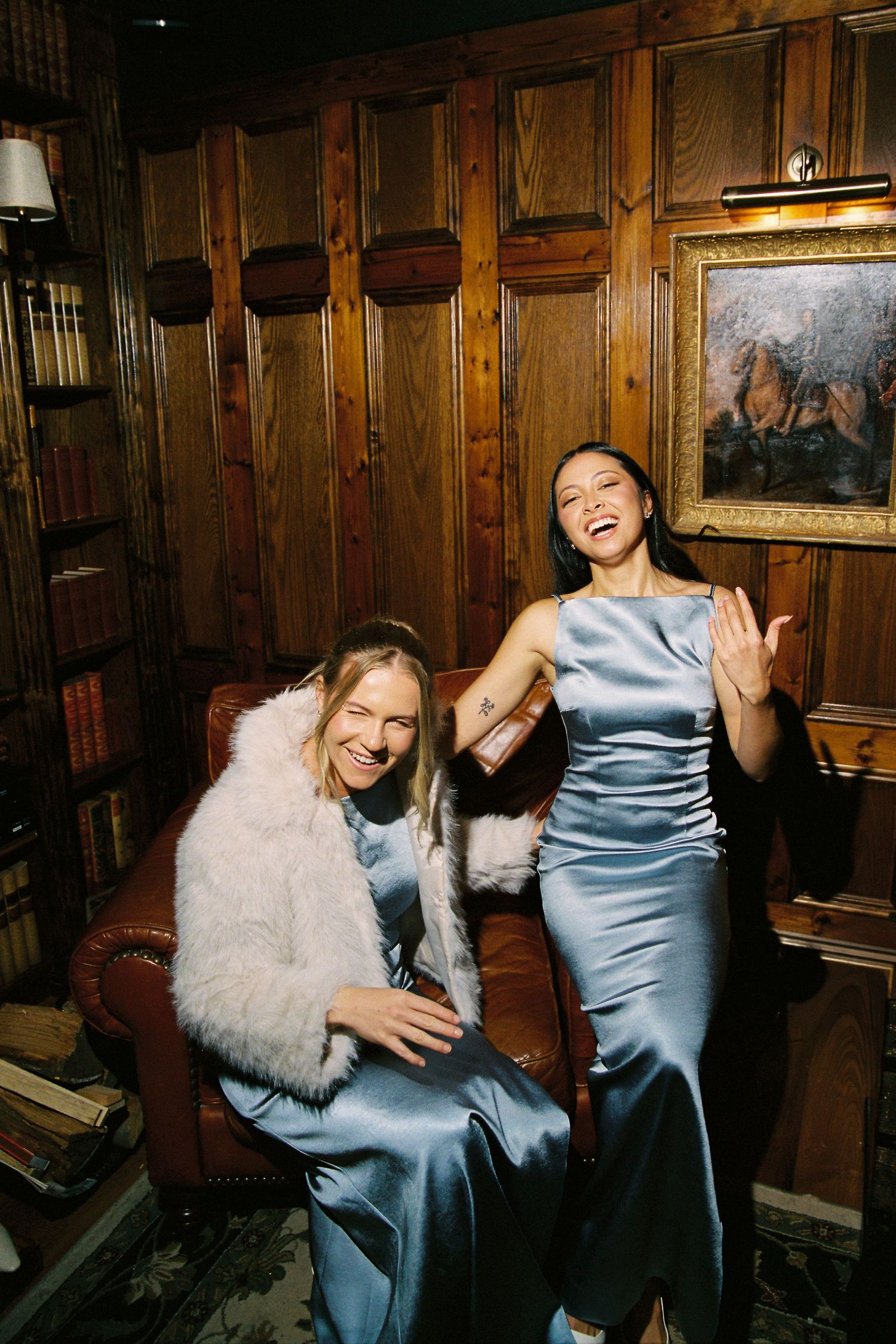 Two bridesmaids in silver satin dresses laughing in wood-paneled library room with leather armchair.