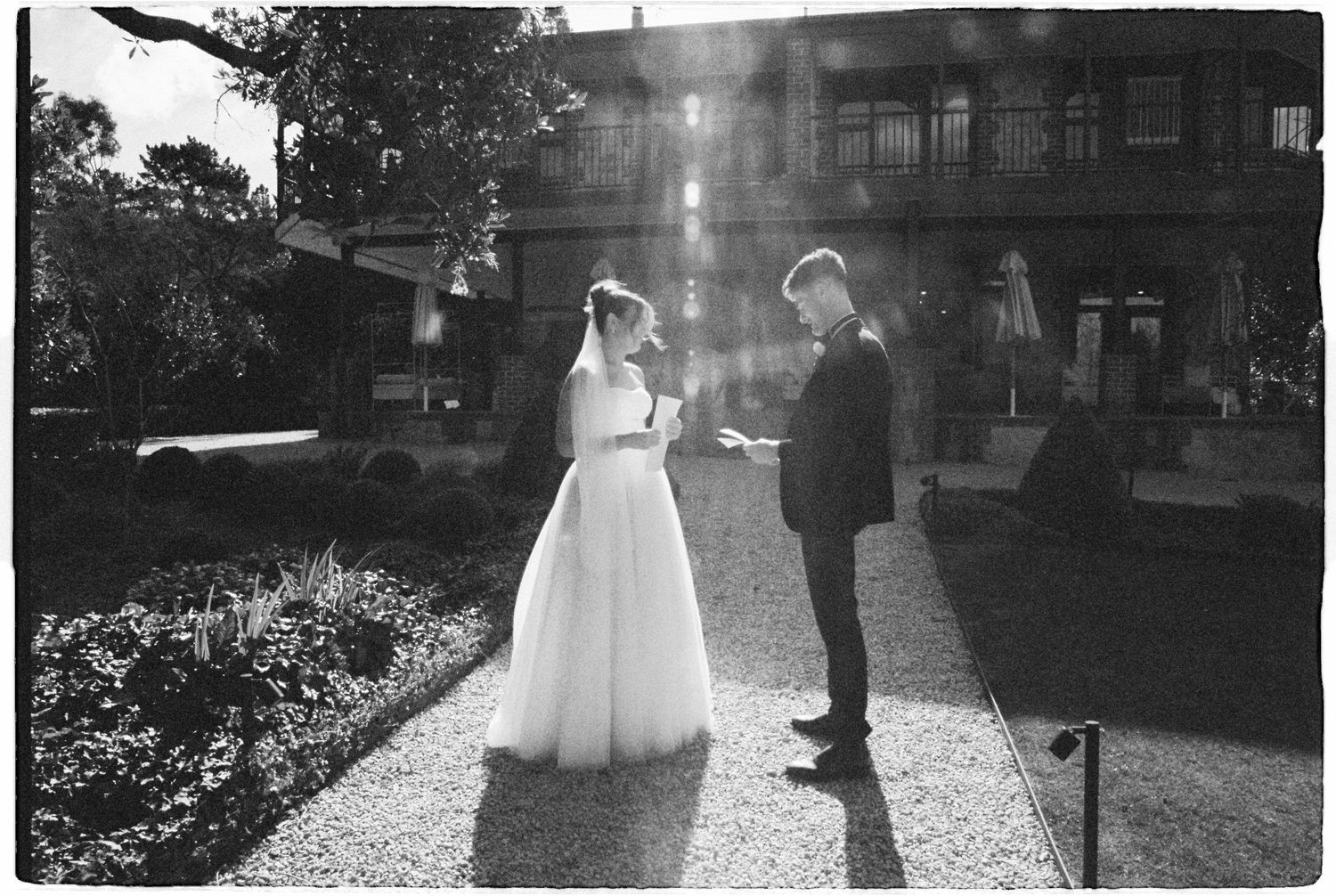 Black and white photo of bride and groom exchanging vows outdoors in a sunlit garden.