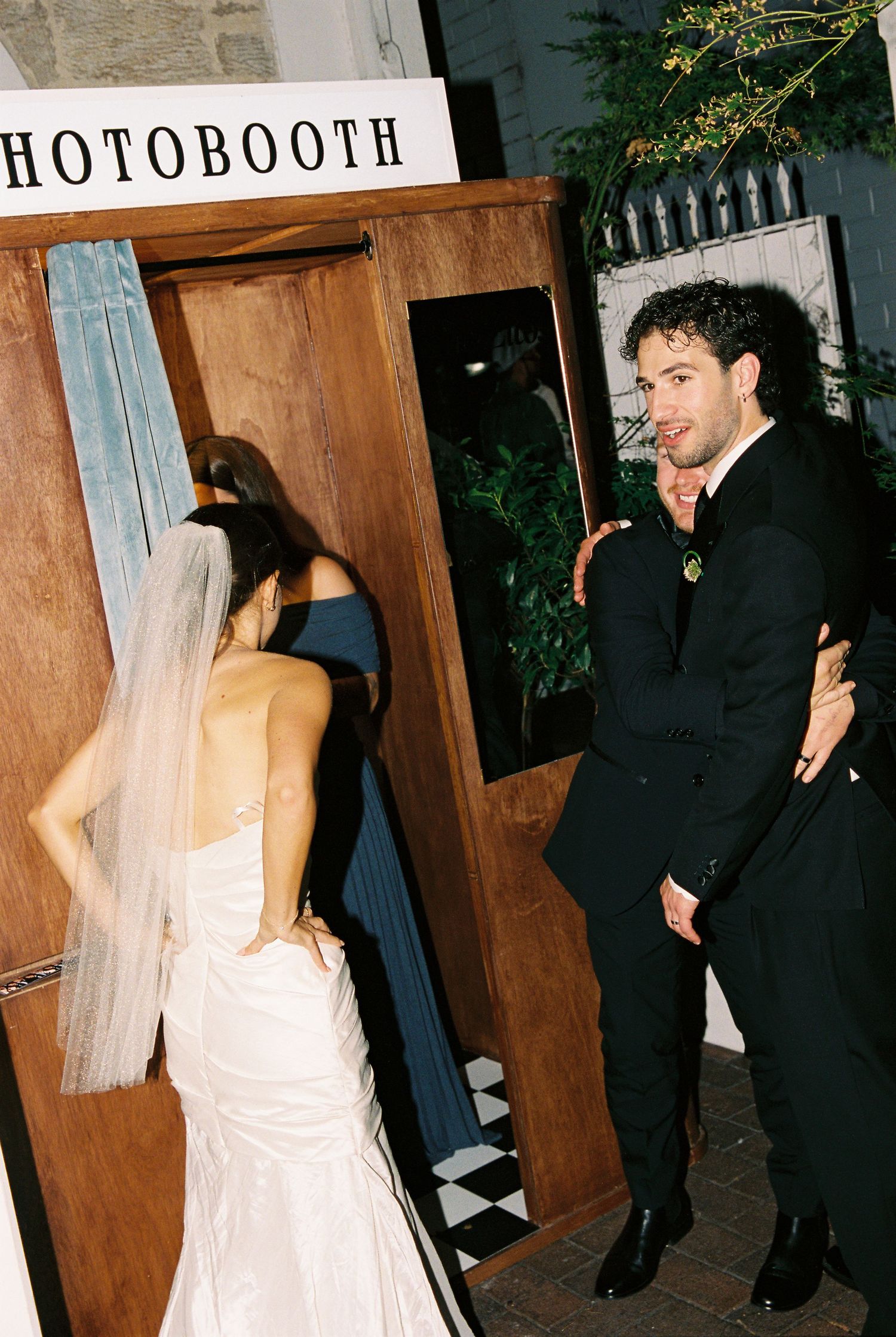 Bride in white gown and veil entering a wooden photo booth with groom in black suit.