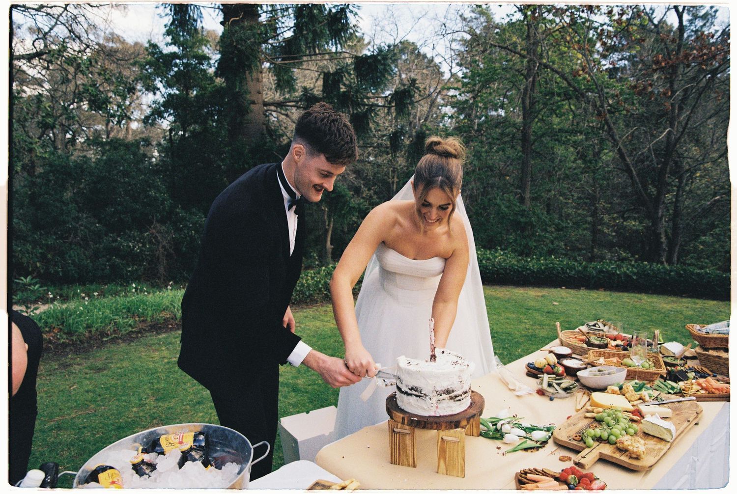 Bride and groom cutting their wedding cake at an outdoor reception table filled with food.