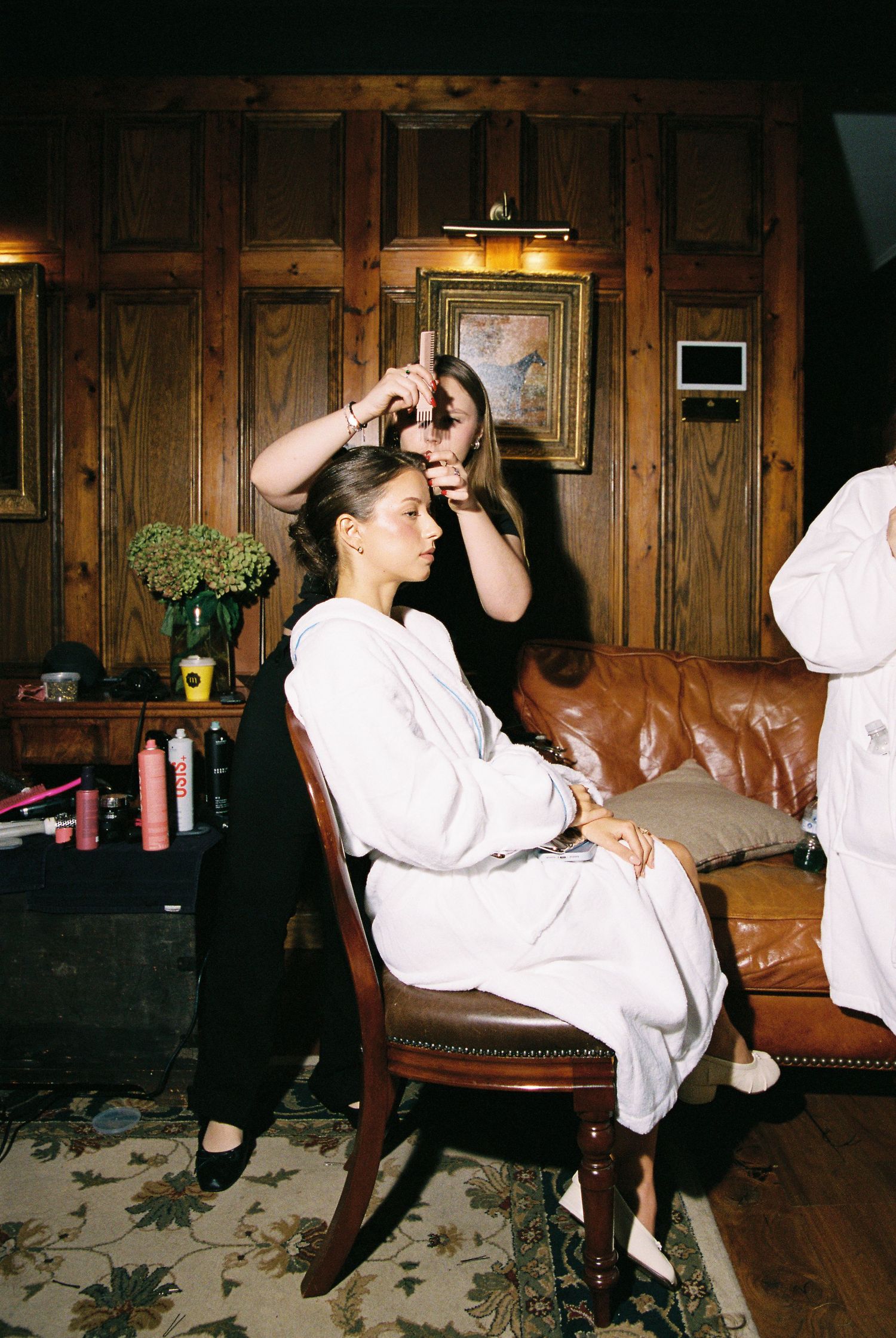 Hairstylist styling bride's hair as she sits in chair in wood-paneled dressing room.