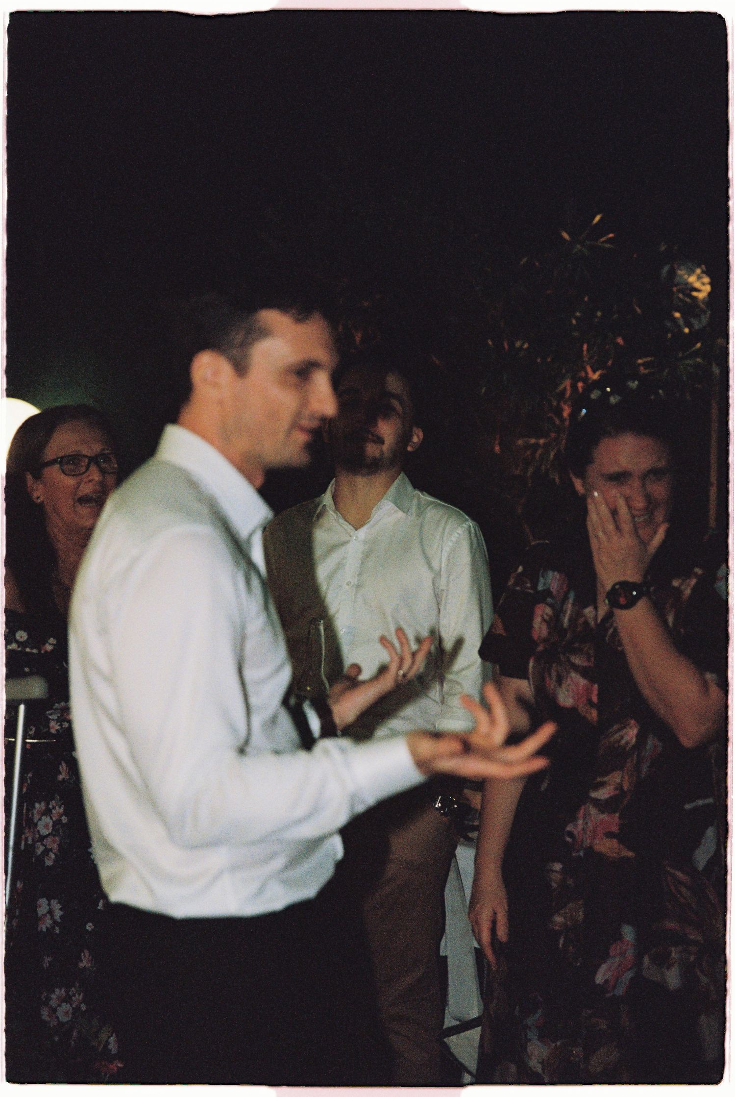 Groom claps while wedding guests laugh and celebrate at evening reception.