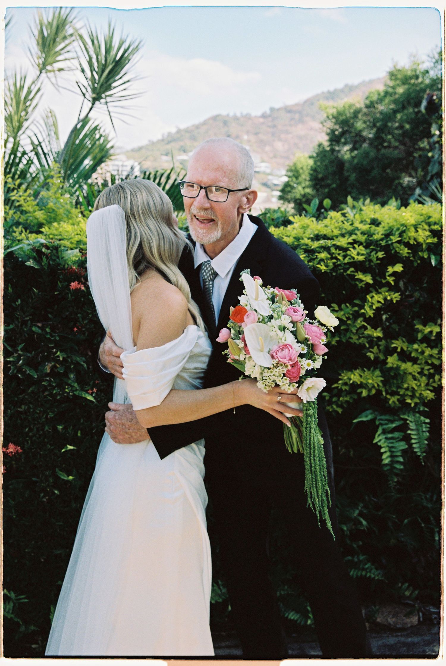 Bride in white gown holding pink bouquet embraces older man in black suit outdoors with lush greenery background.
