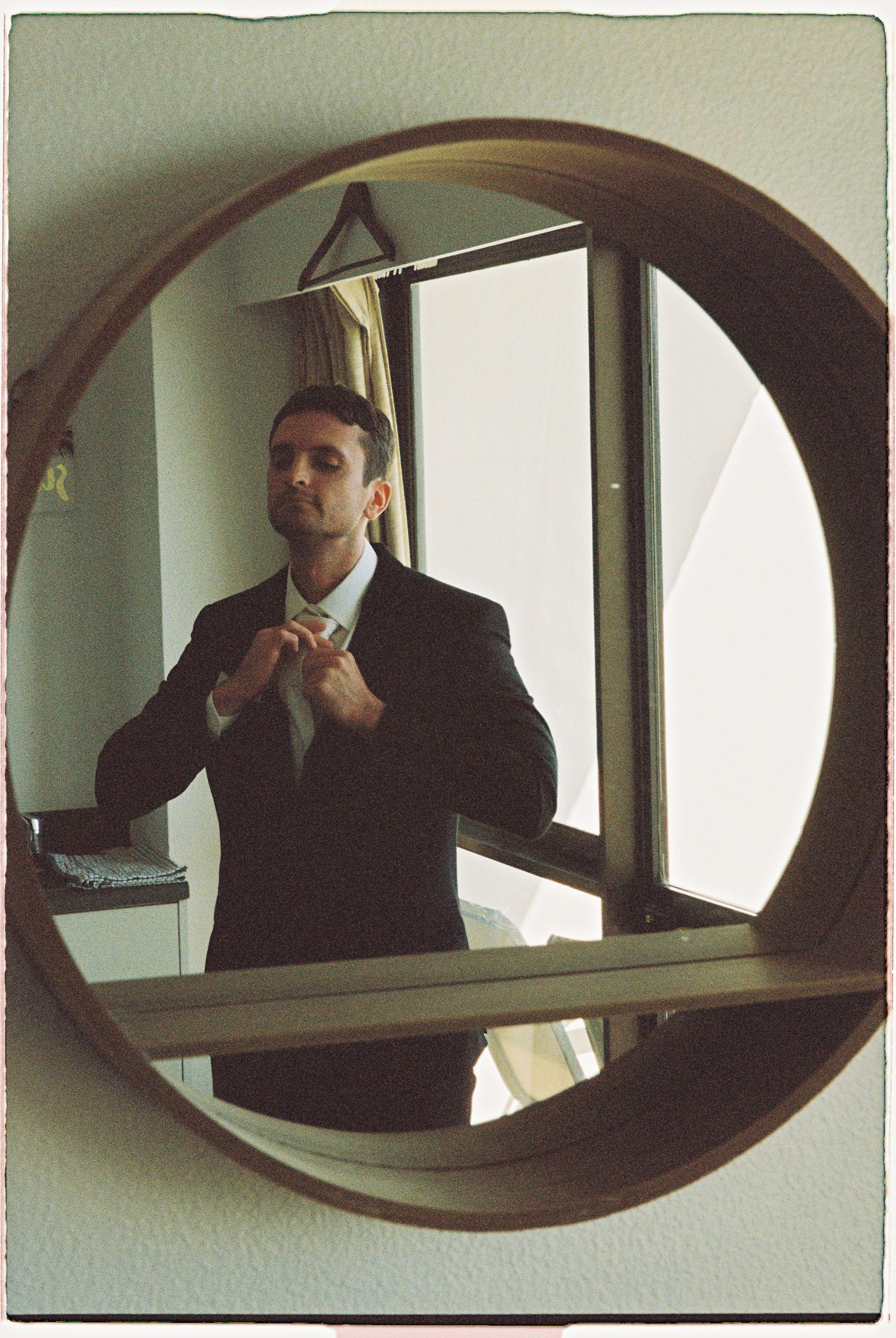 Groom in dark suit adjusting tie reflected in round wooden-framed mirror in hotel room.