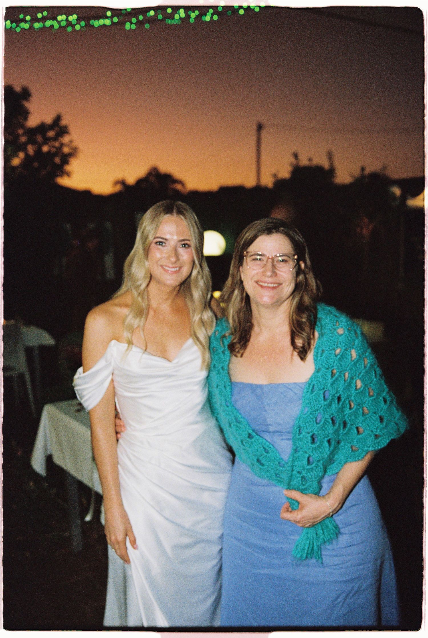 Bride in white off-shoulder gown poses with guest in blue dress and teal shawl at outdoor sunset wedding.