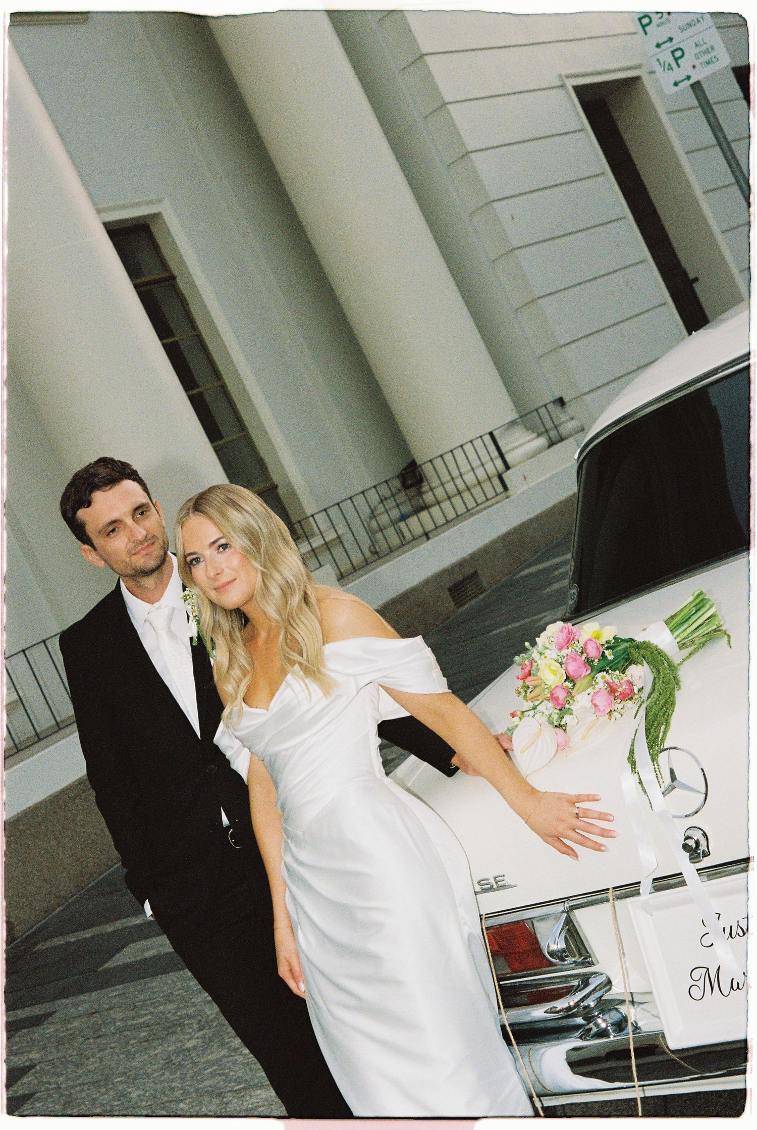 Bride in white off-shoulder gown and groom in tuxedo pose beside a decorated Mercedes wedding car outside a grand building.