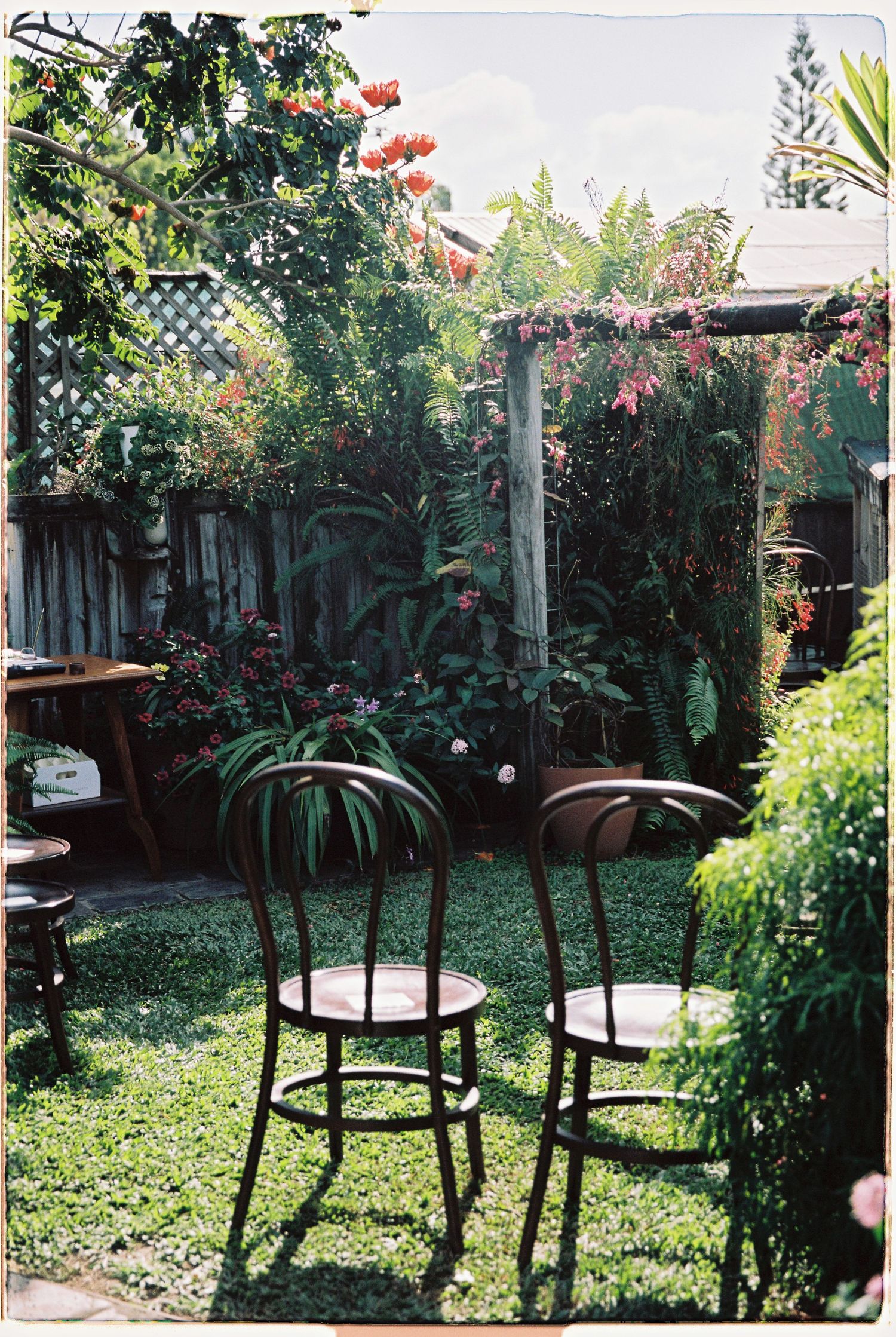 Rustic garden setting with vintage bistro chairs on green lawn surrounded by colorful flowering plants.