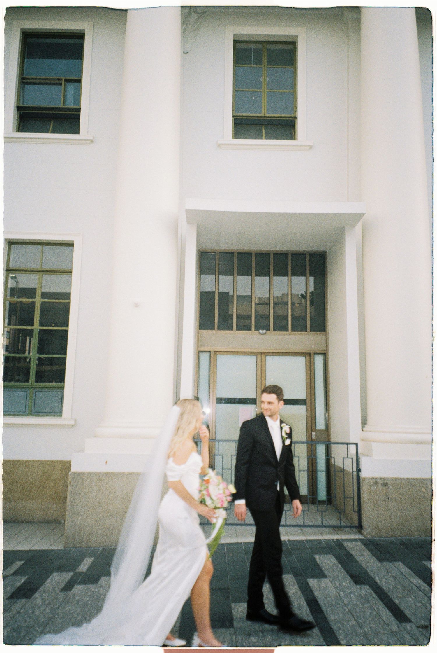 Elegant bride in white gown with veil walks with tuxedo groom outside a grand white building.