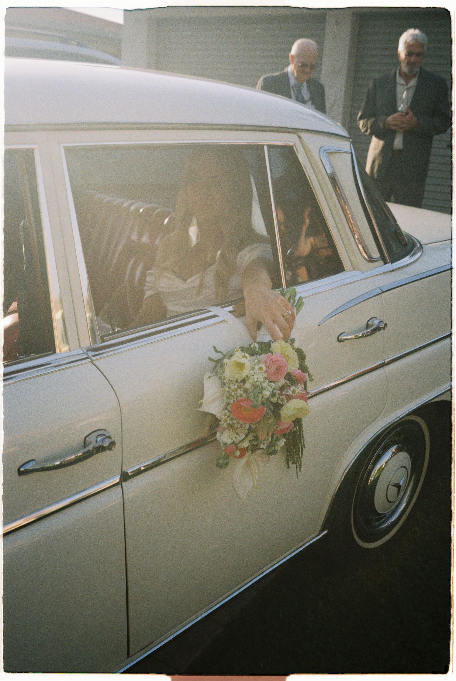 Bride holding colorful bouquet through window of classic vintage white wedding car.