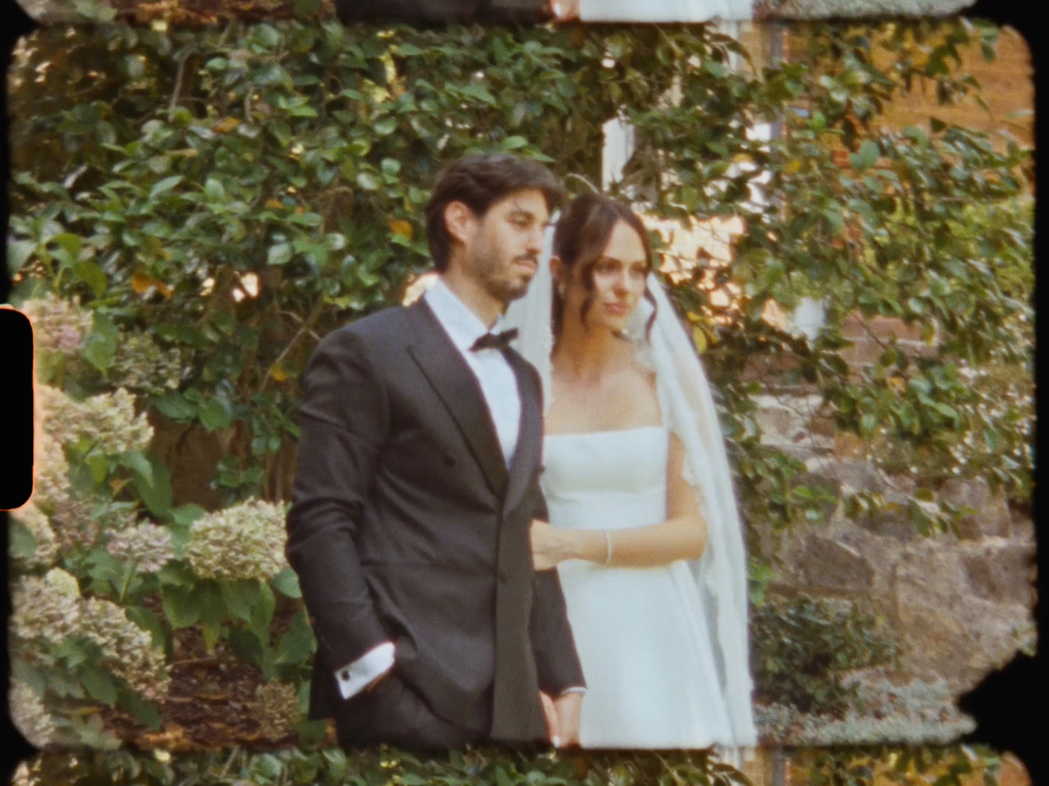 Bride and groom in elegant wedding attire posing together surrounded by lush green garden foliage.