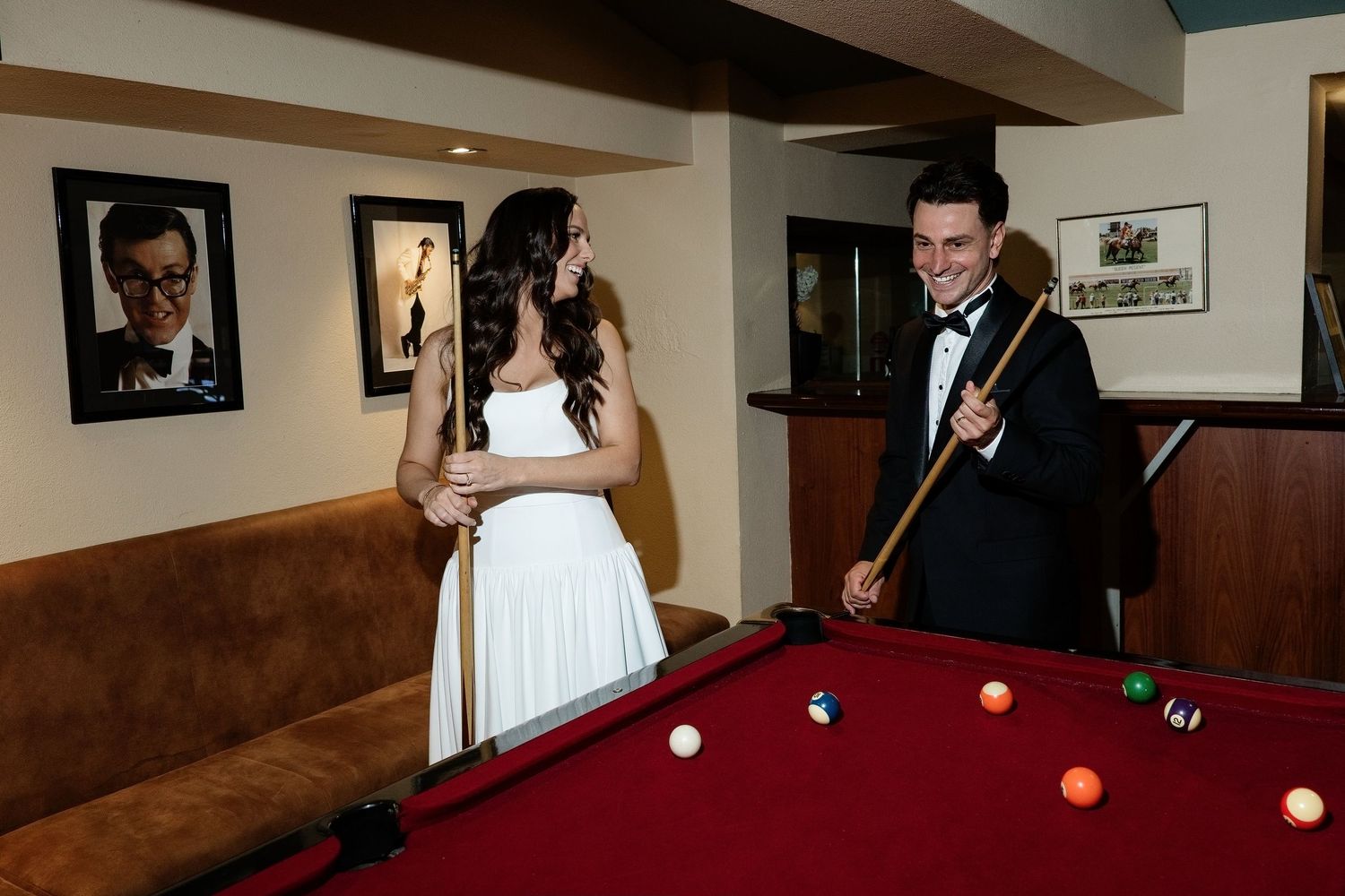 Bride in white dress and groom in tuxedo standing together beside a red pool table with billiard balls.