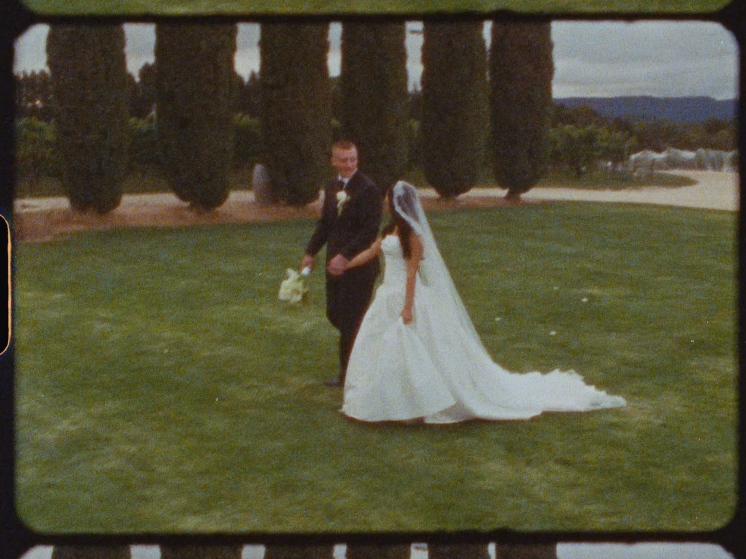 Bride in flowing white gown with veil walking with groom on green lawn surrounded by cypress trees.