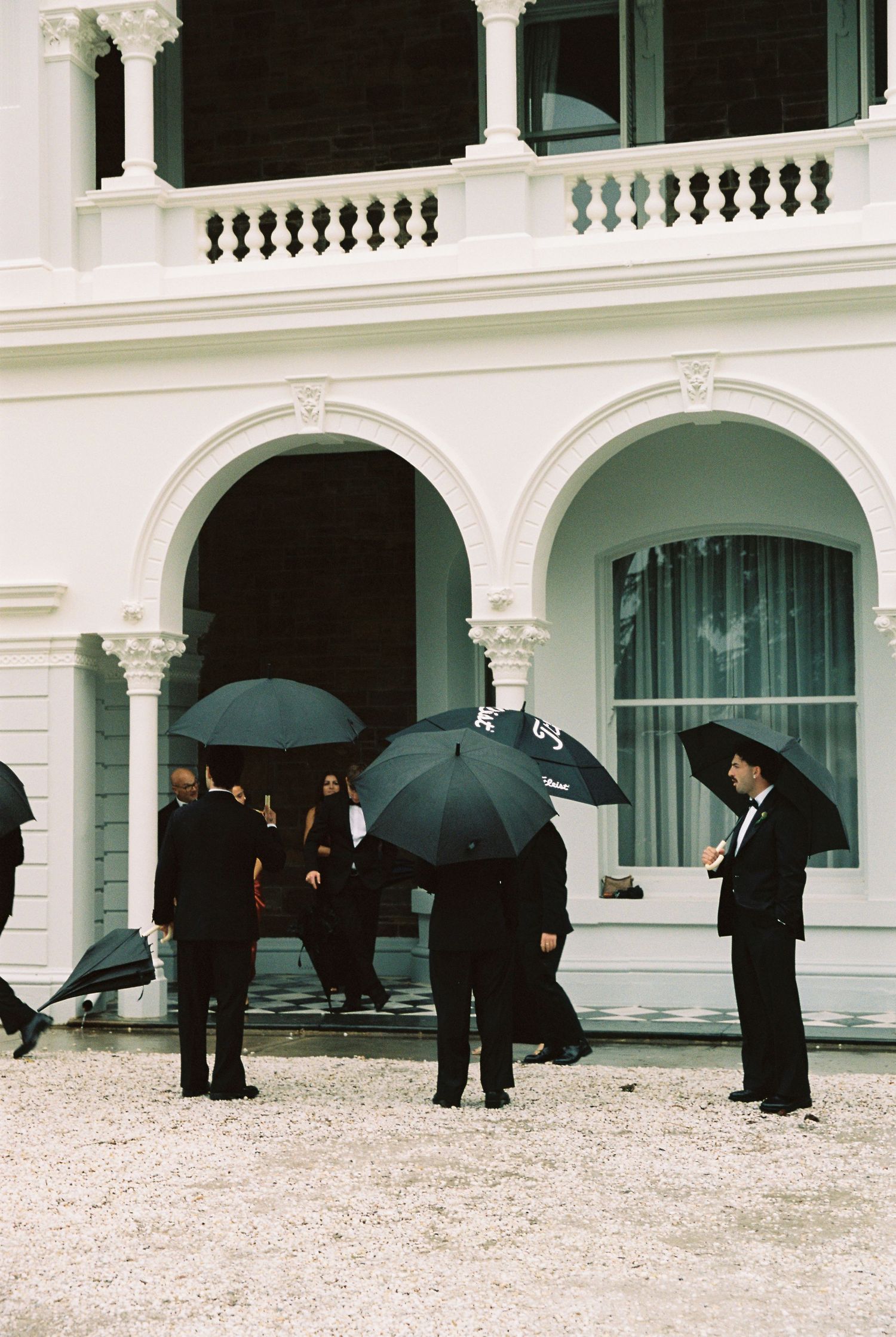 Guests in formal black attire holding black umbrellas outside a white colonial building with arched entrances.