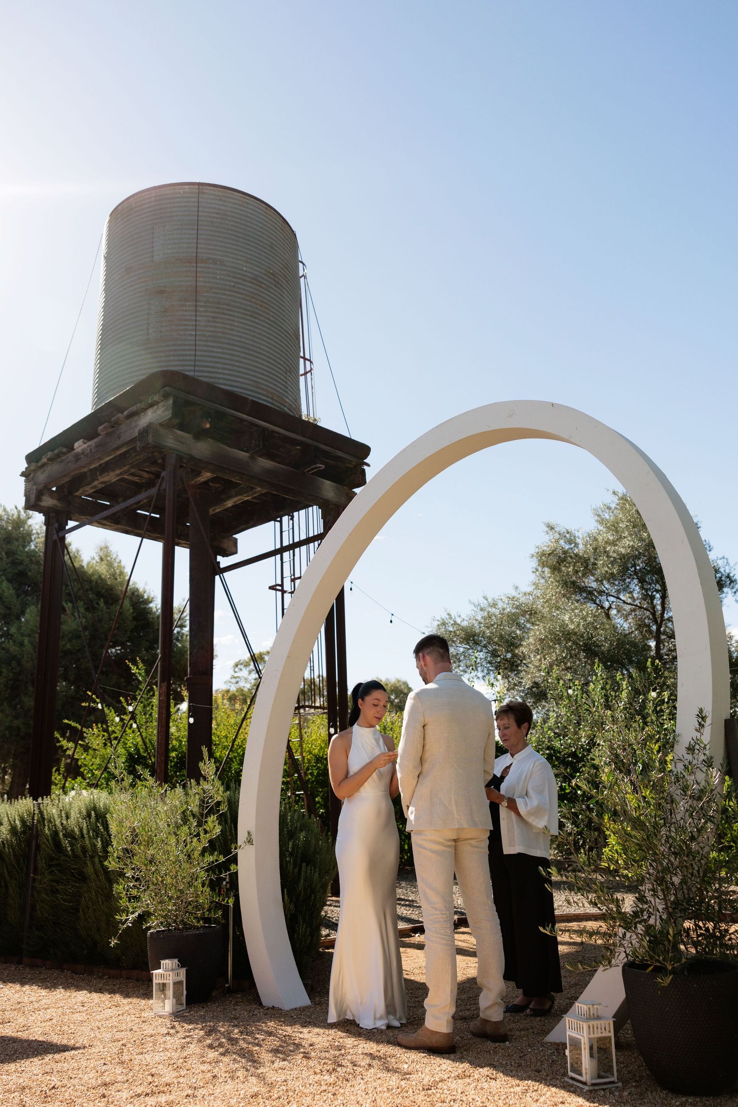 Couple exchanging vows under white arch with rustic water tower backdrop on sunny wedding day.