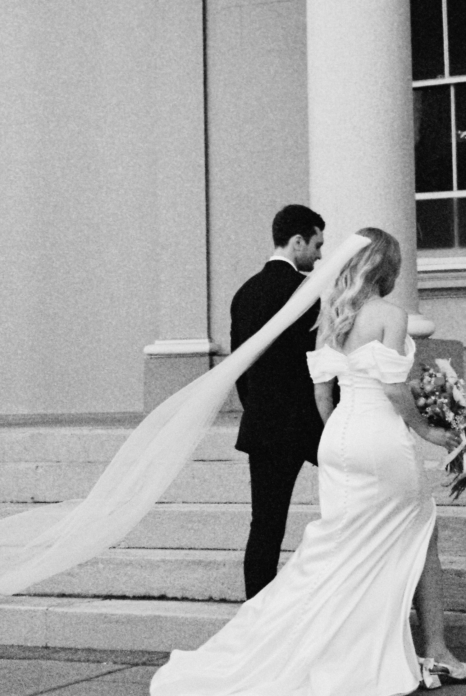 Black and white photo of bride in fitted gown with long veil walking with groom up steps.