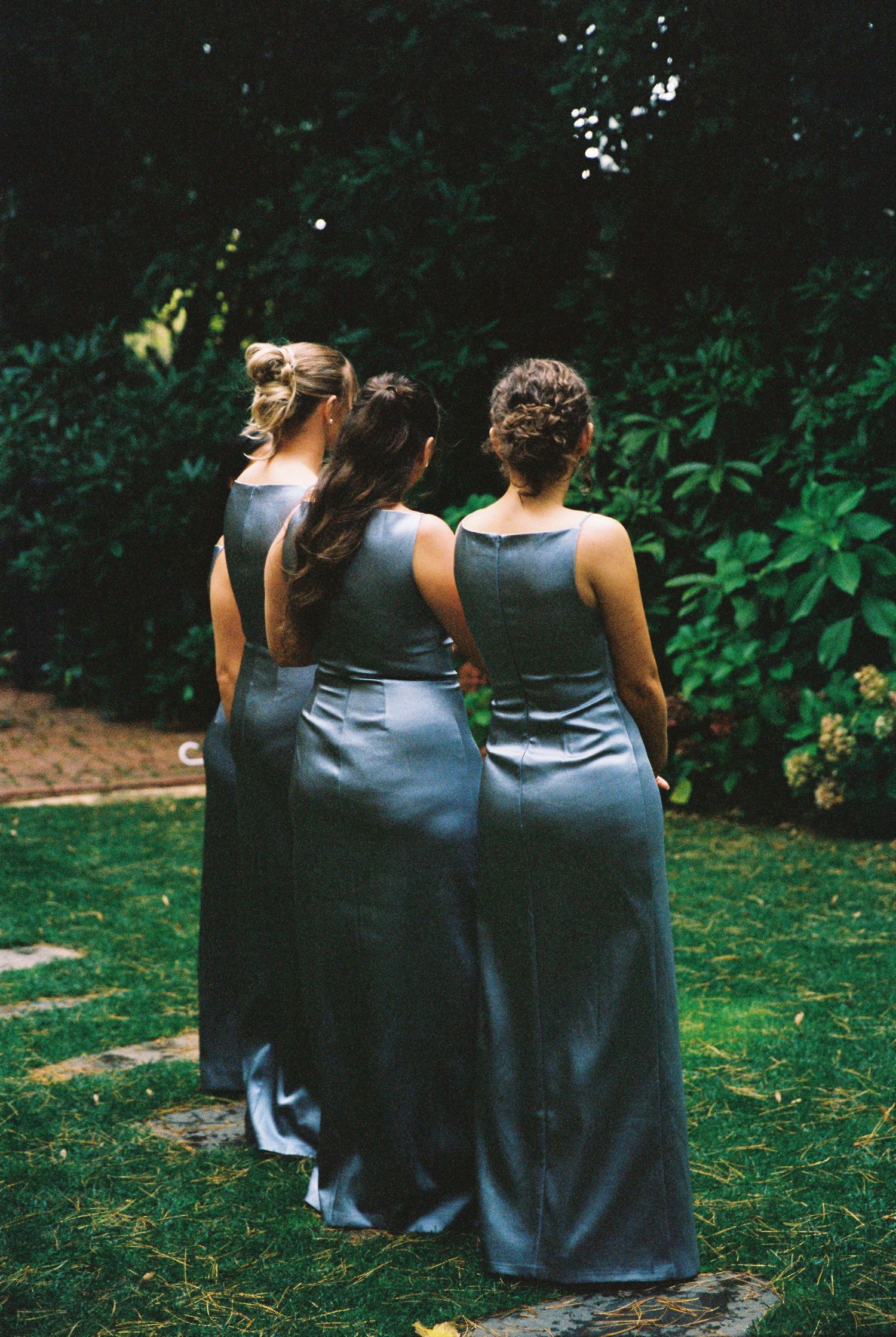 Three bridesmaids in matching slate blue satin gowns standing with backs to camera in lush garden.