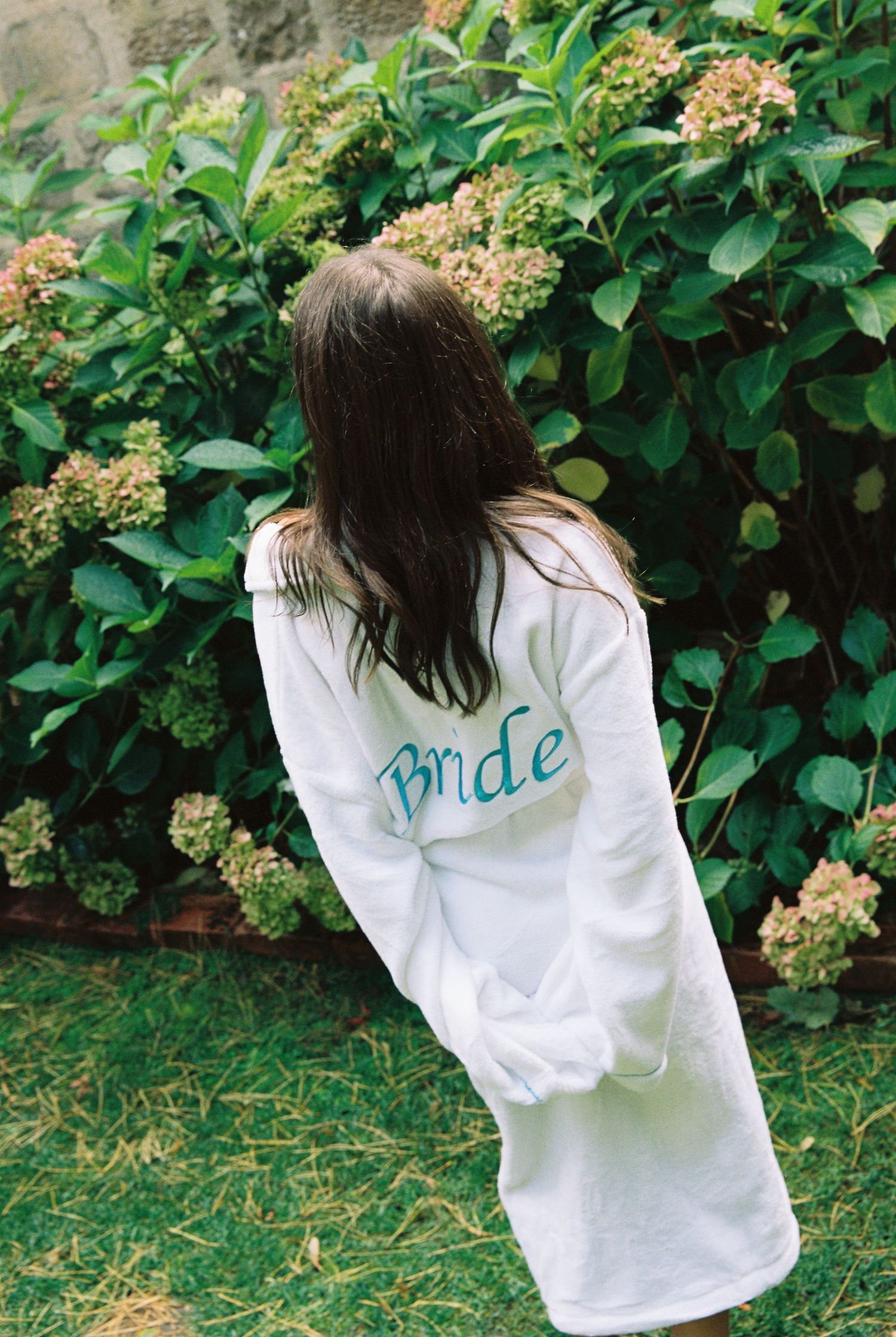 Bride wearing white robe with teal Bride embroidery standing near green hydrangea bushes outdoors.