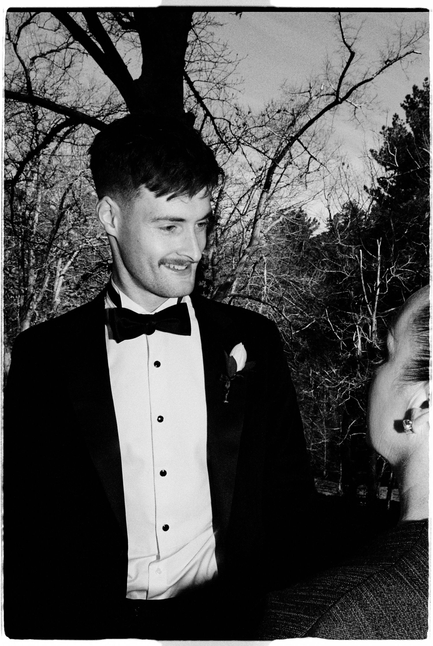 Man in tuxedo with bow tie smiling outdoors at winter wedding ceremony with bare trees in background.