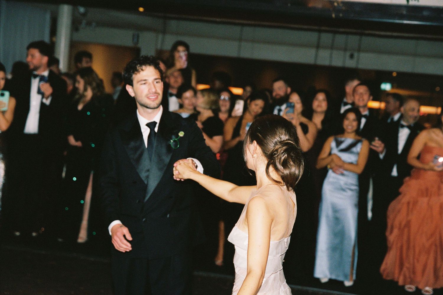 Groom in black tuxedo sharing first dance with bride before applauding wedding guests.