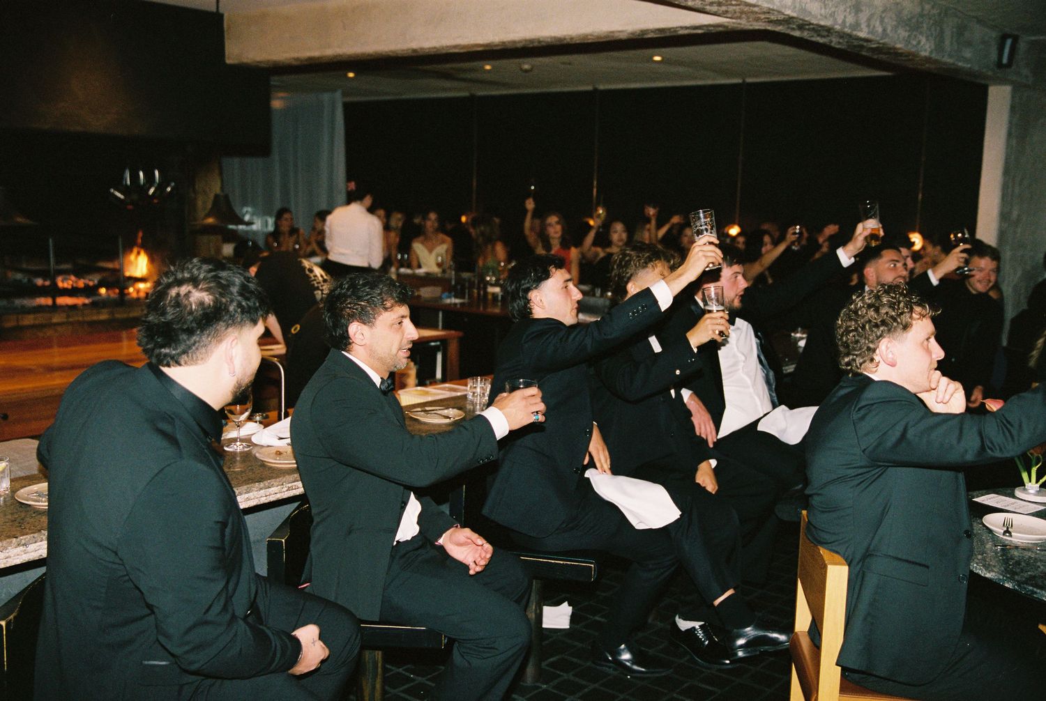 Guests in formal black attire raising glasses in a toast at a wedding reception dinner.