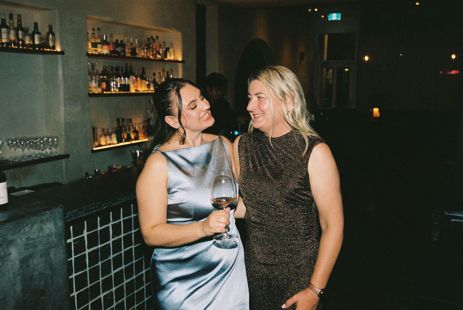Two women smiling together at a bar, one in a silver dress holding red wine, at an upscale venue.