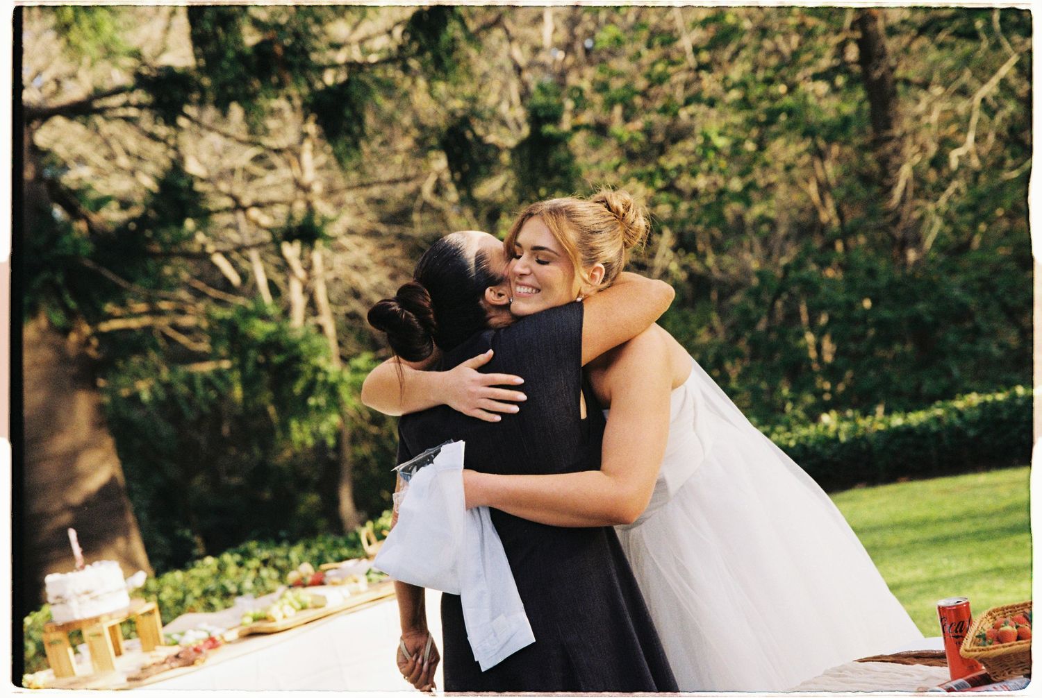 Bride in white gown hugging a guest at an outdoor wedding reception surrounded by greenery.