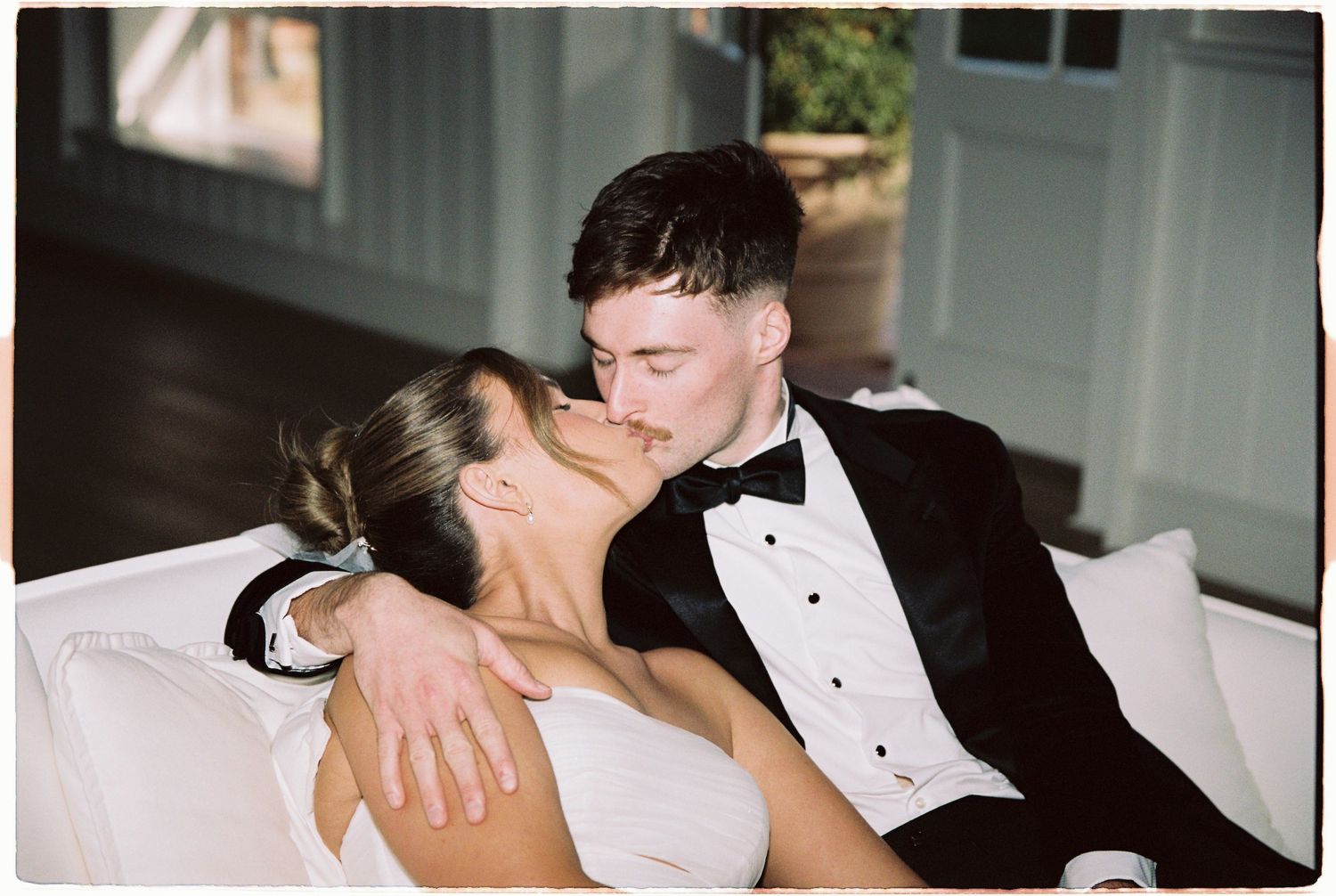 Bride and groom sharing a kiss on a white couch at their wedding reception.