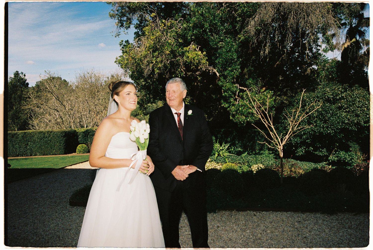 Bride in white gown holding bouquet stands with older man in dark suit at outdoor wedding venue.