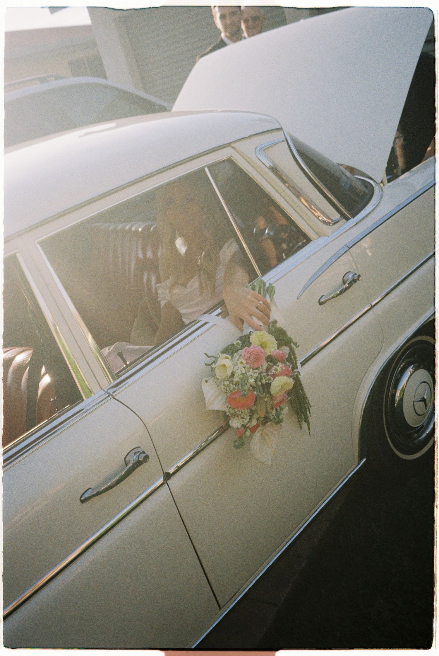 Bride holding colorful floral bouquet through window of vintage white classic wedding getaway car.