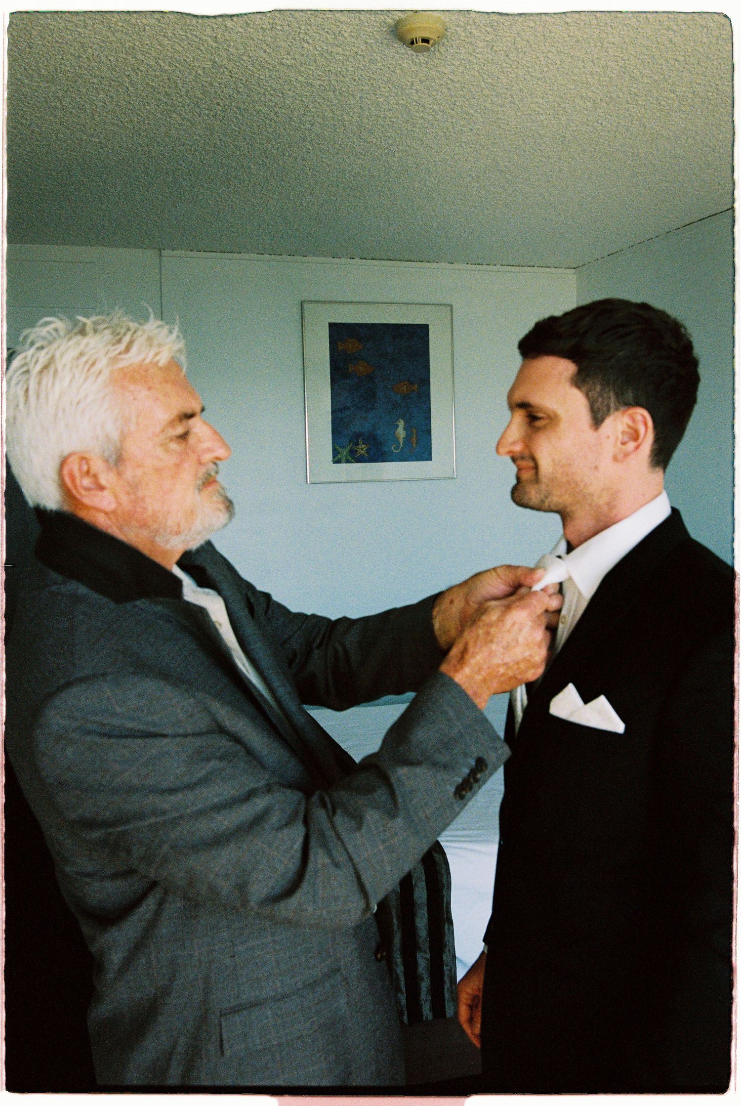 Older man with white hair helping groom adjust his bow tie before wedding ceremony indoors.