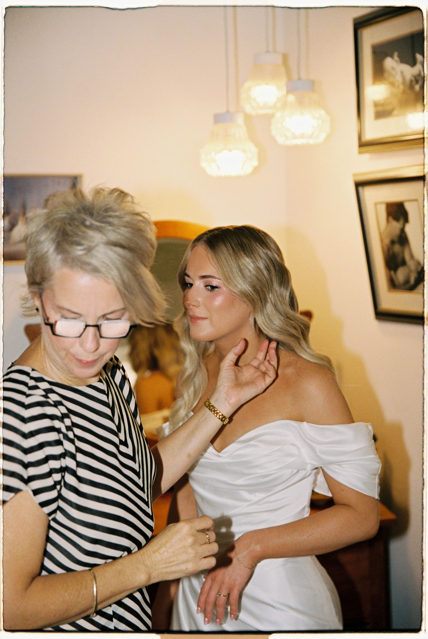 Older woman helping blonde bride with necklace in warmly lit room before wedding ceremony.