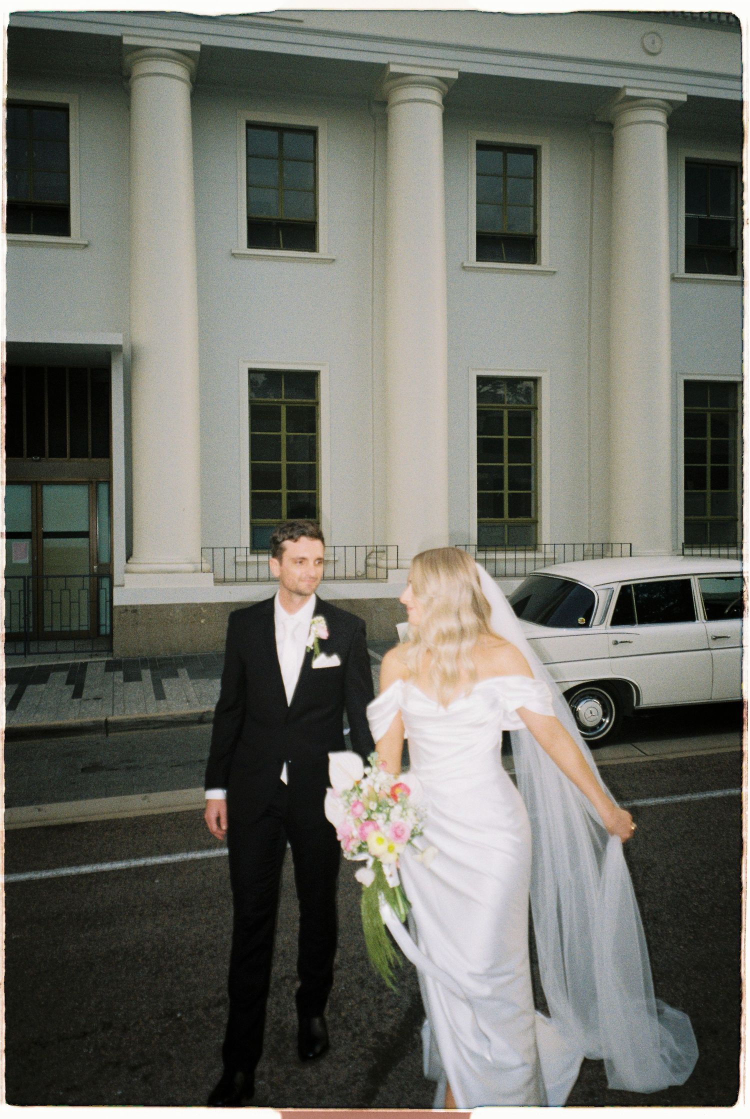 Bride and groom walk near white classic car outside grand white building on wedding day.