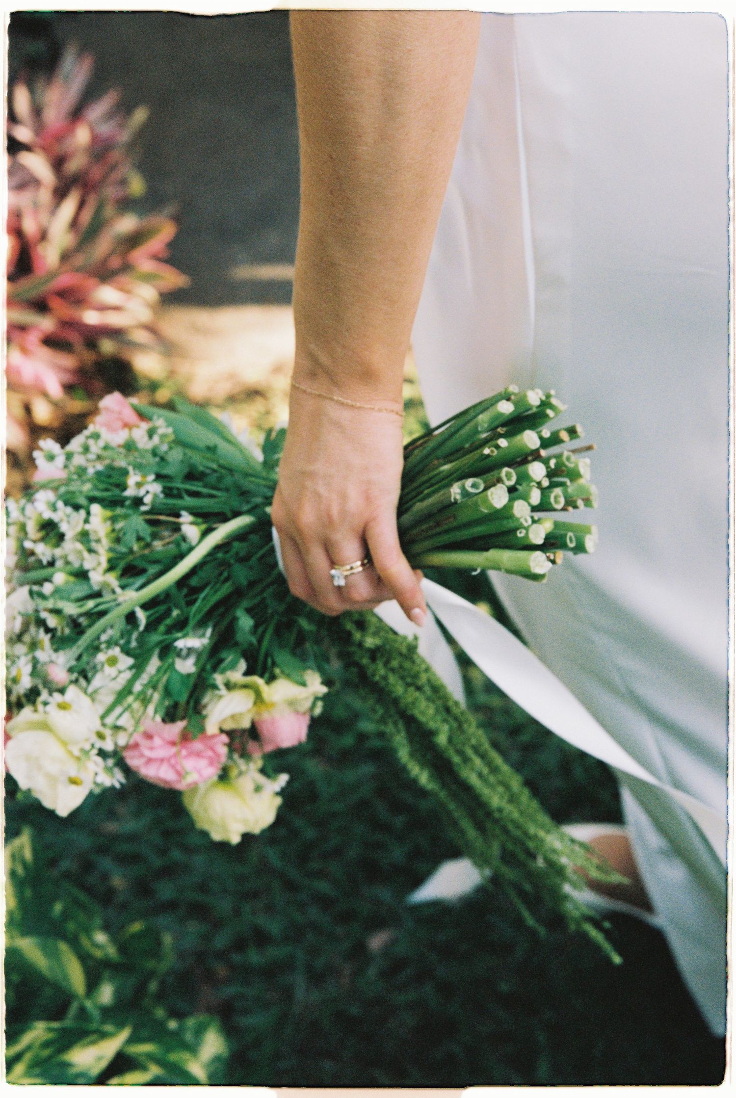 Close-up of bride holding green stemmed wedding bouquet with pink and white flowers, wearing stacked rings.